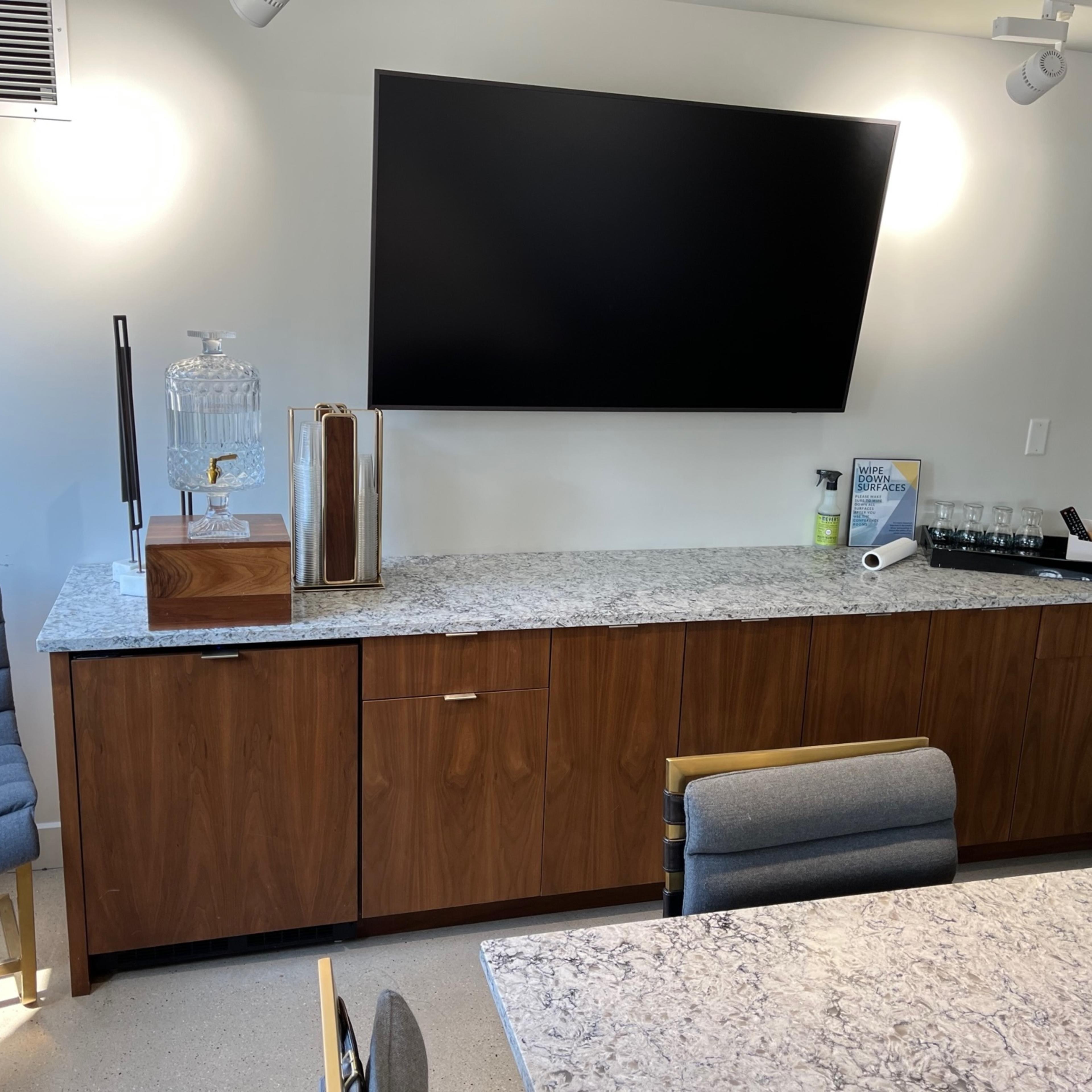 A modern conference room with a wooden cabinet below a wall-mounted television, a stone countertop, and a seating area featuring a gray chair.