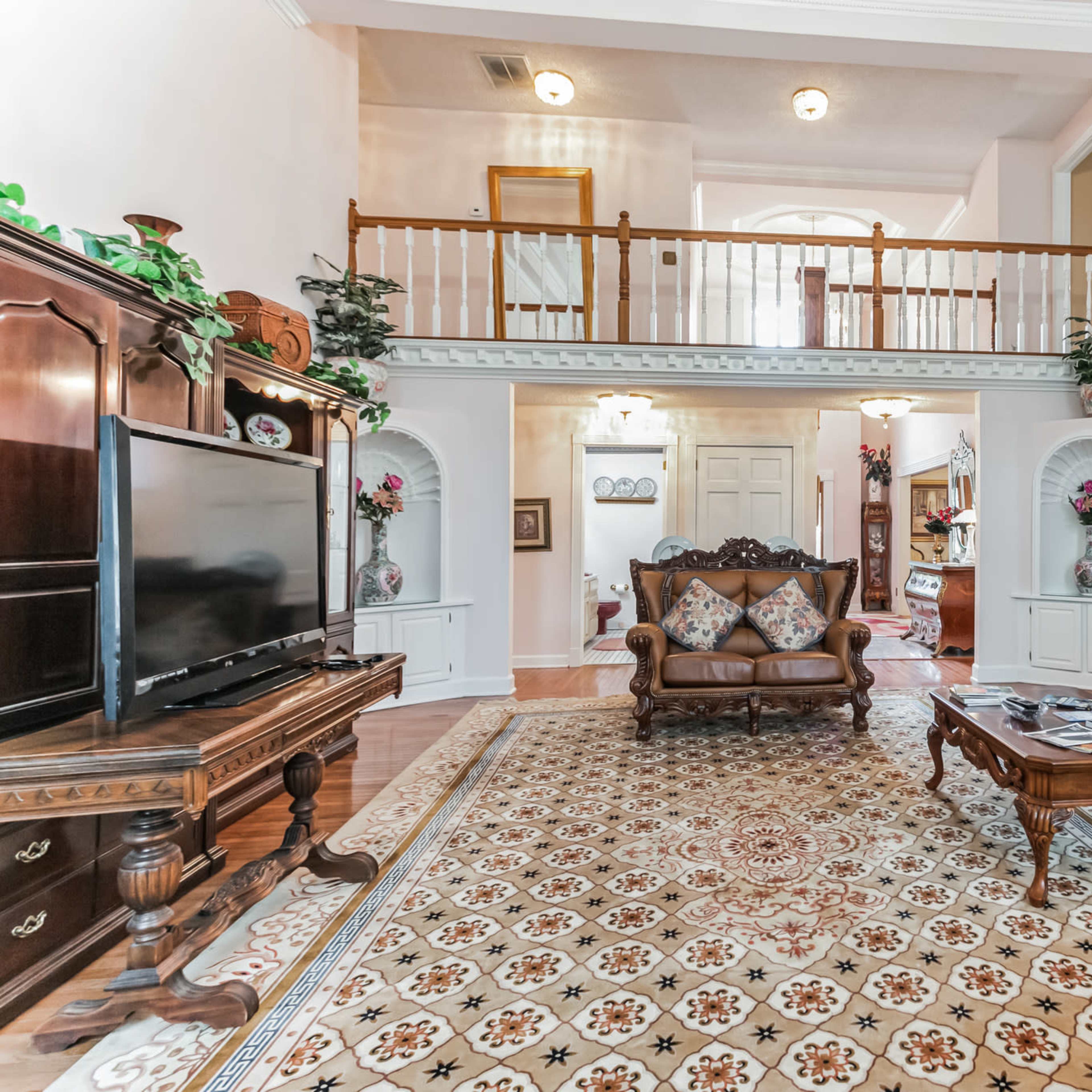 A spacious living room with wooden flooring, decorative furniture, a large television, and a balcony overlooking the area.