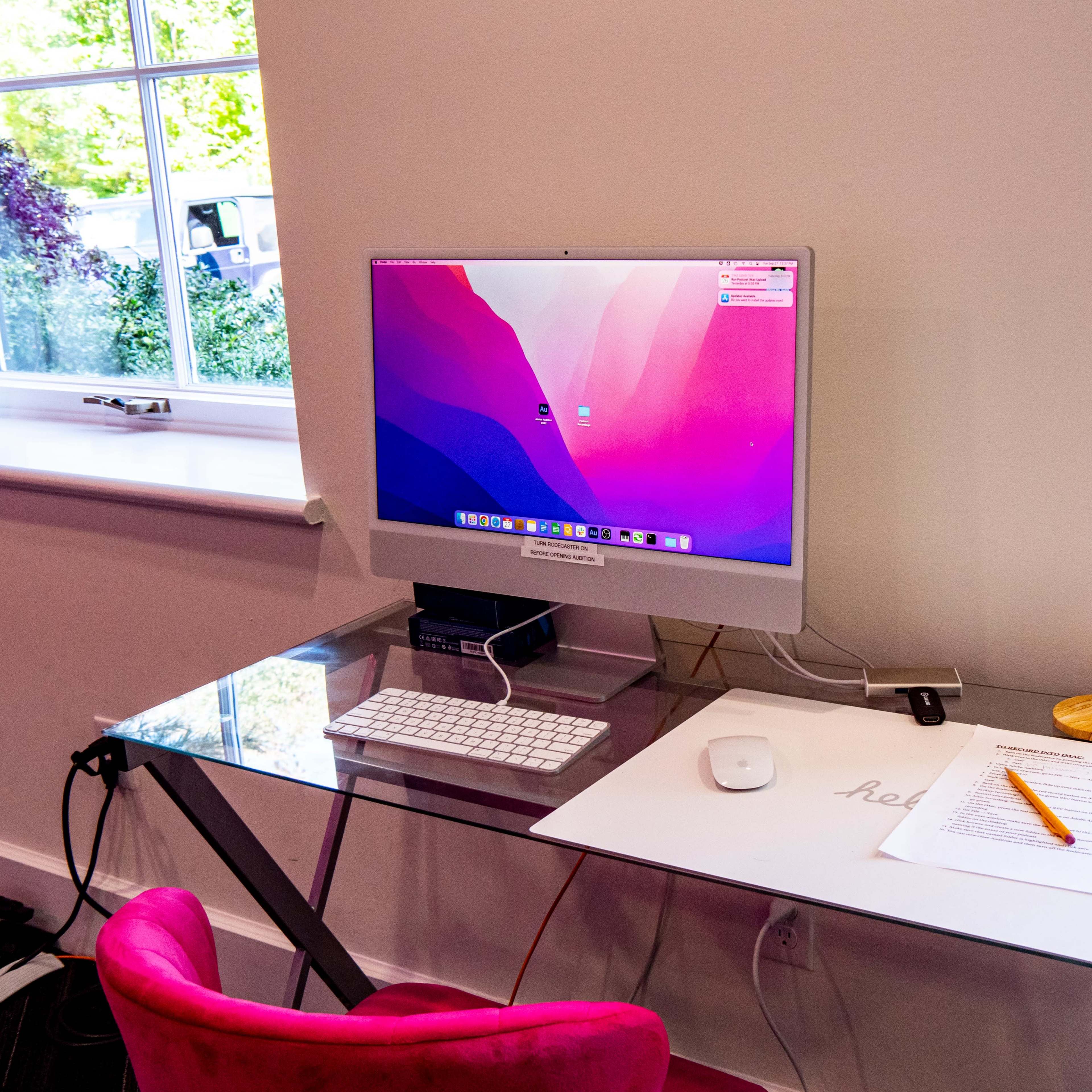 A modern workspace features an Apple computer on a glass desk with a keyboard, mouse, and a document alongside an orange pen.