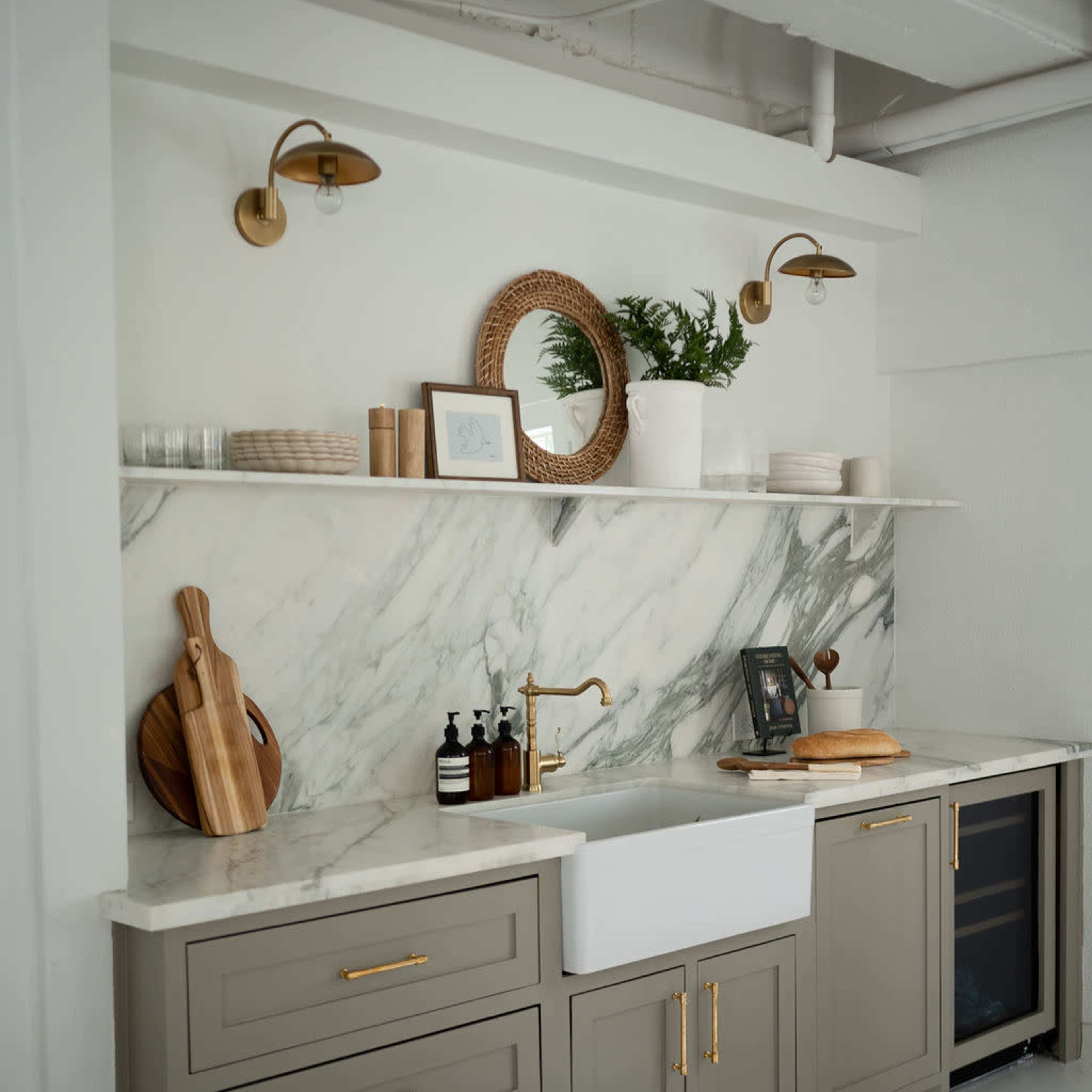 A modern kitchen features a marble backsplash, a farmhouse sink, gray cabinetry, and wall-mounted sconces.