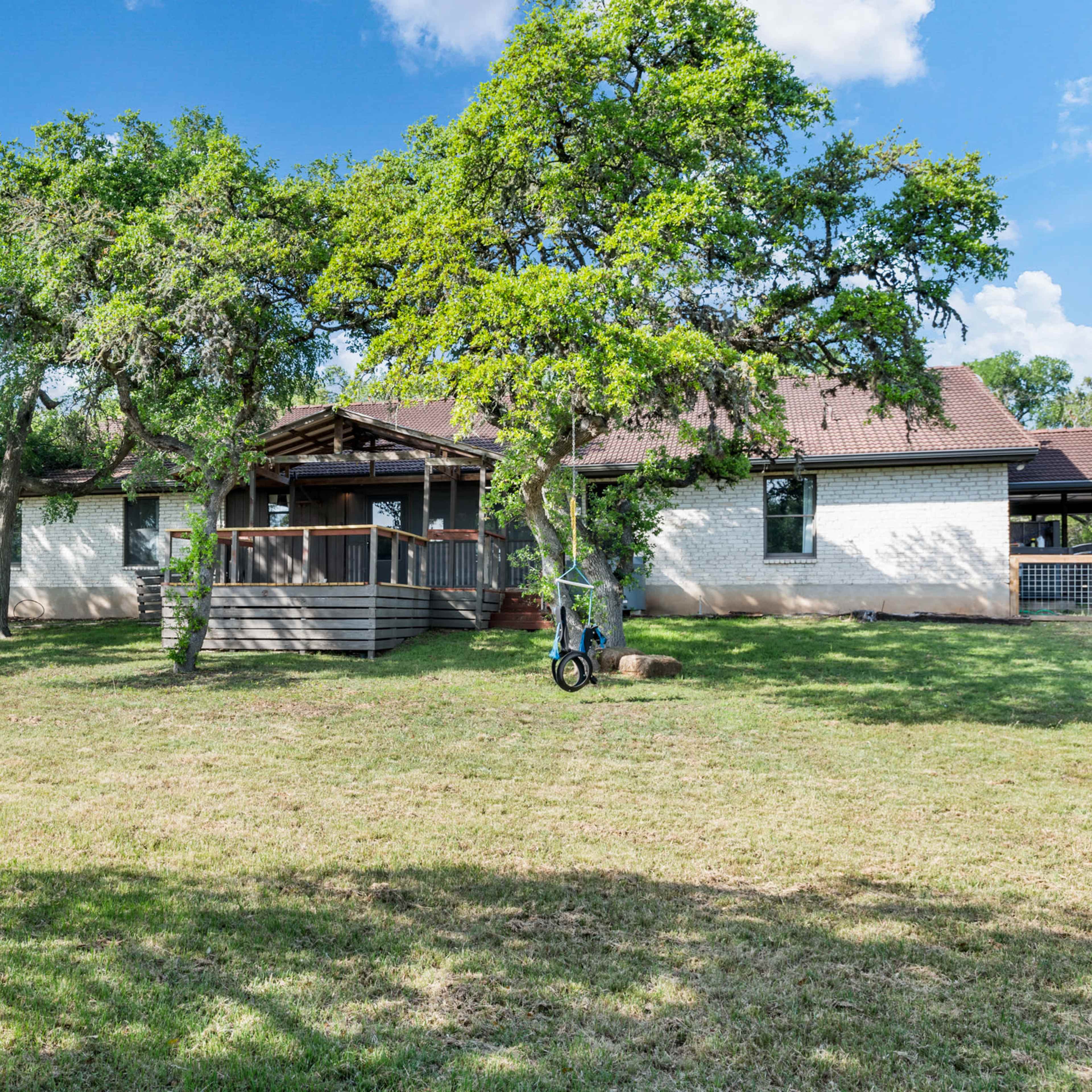 A single-story house with a wrap-around deck is surrounded by several trees and a grassy lawn.