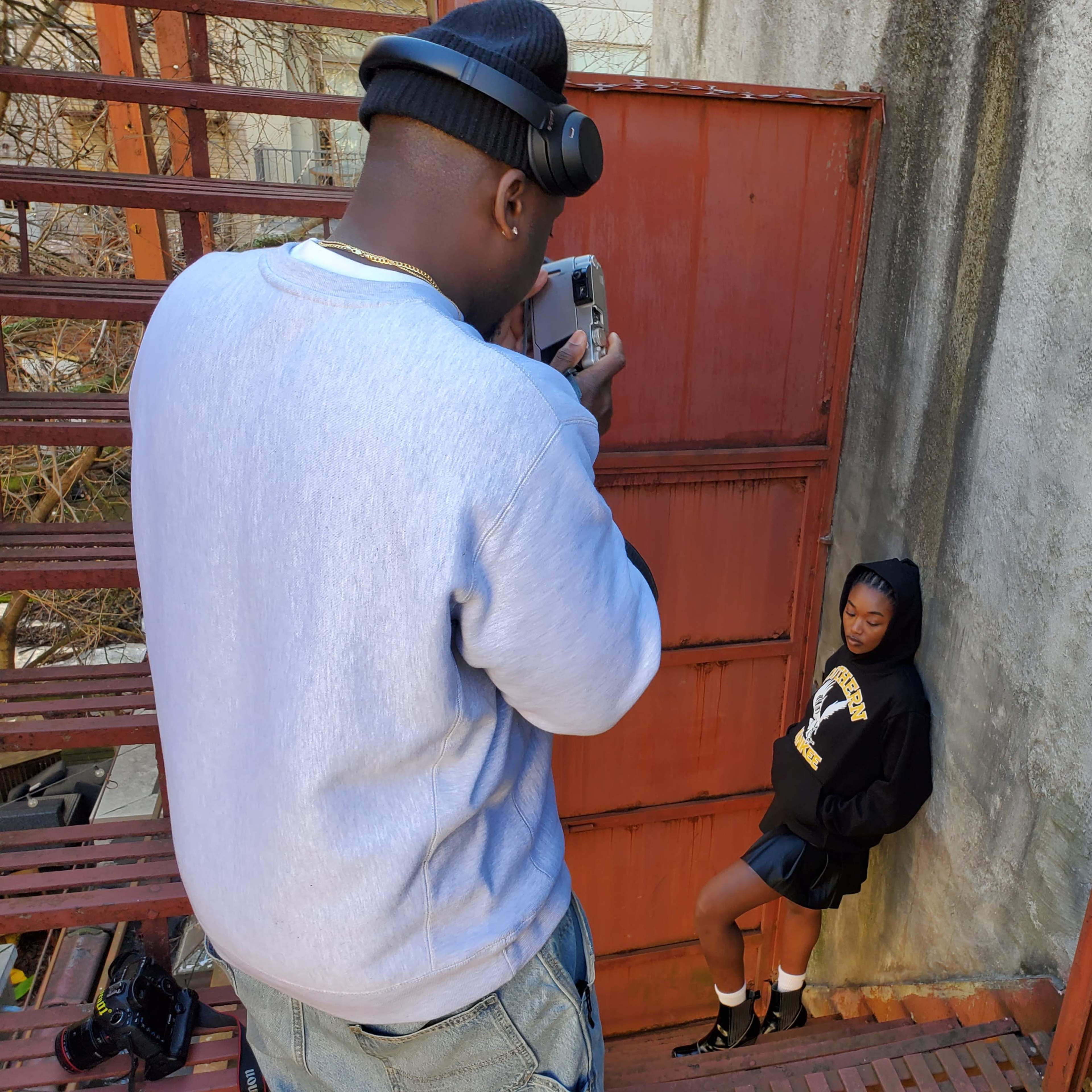 A photographer takes pictures of a model posing in a hoodie and skirt next to a rusted metal wall.