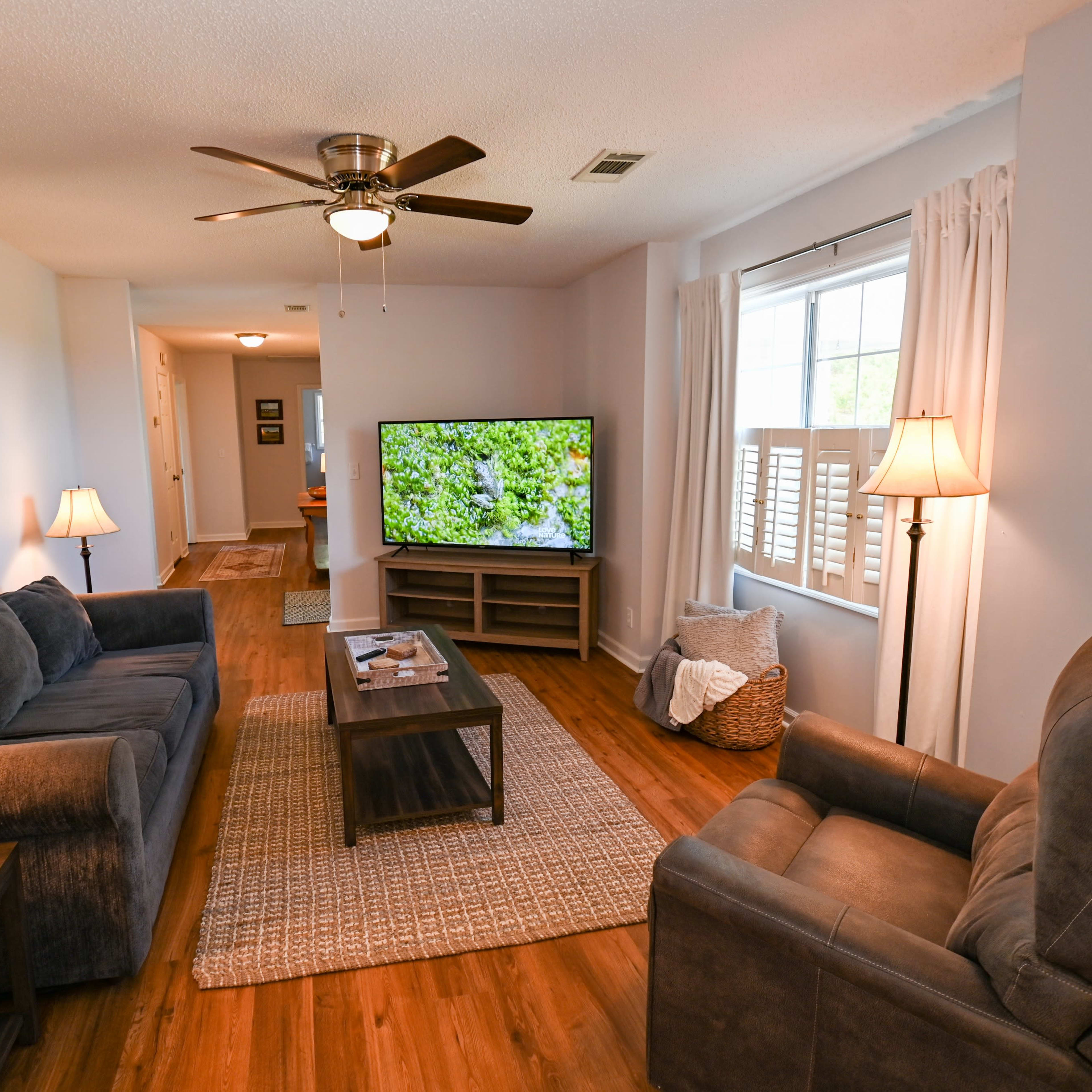 The living room features a sectional sofa, a recliner, a coffee table, and a large television, with wooden floors and light-colored walls.