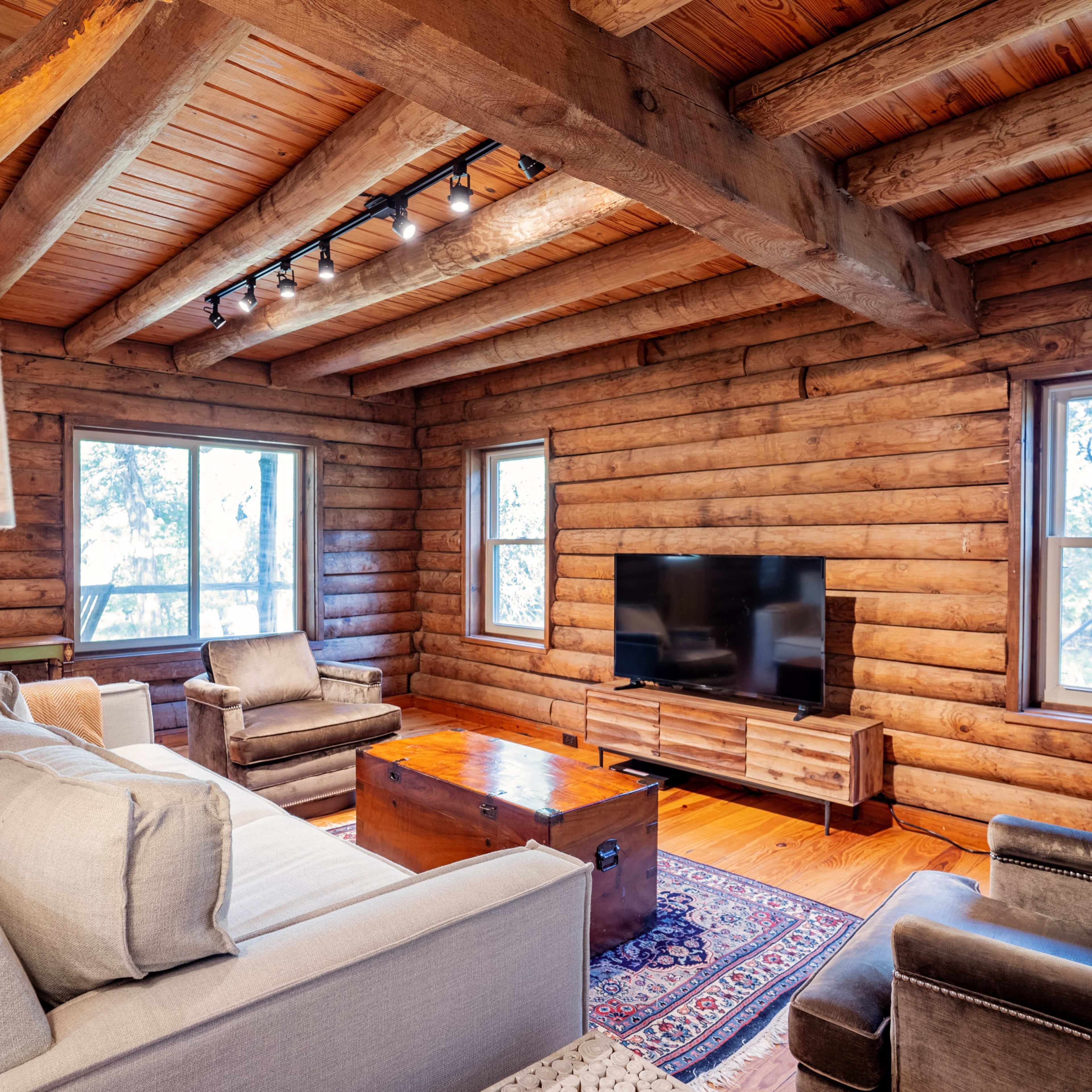 The cozy living room features log walls, wooden beams, a television on a wooden stand, and comfortable seating arranged around a coffee table.