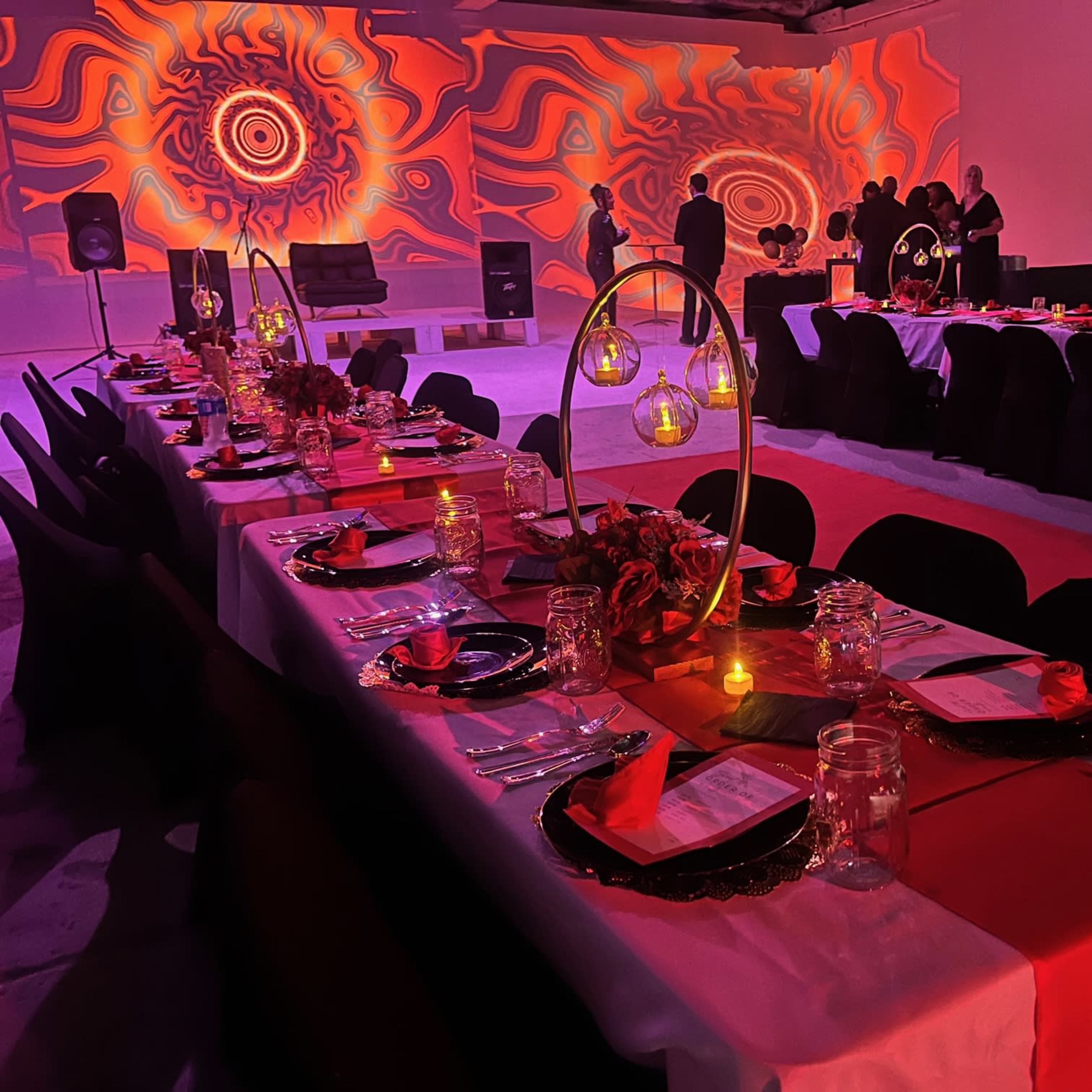 A banquet setup with elegantly arranged tables featuring red and white tablecloths, decorated with centerpieces, candles, and a colorful backdrop.