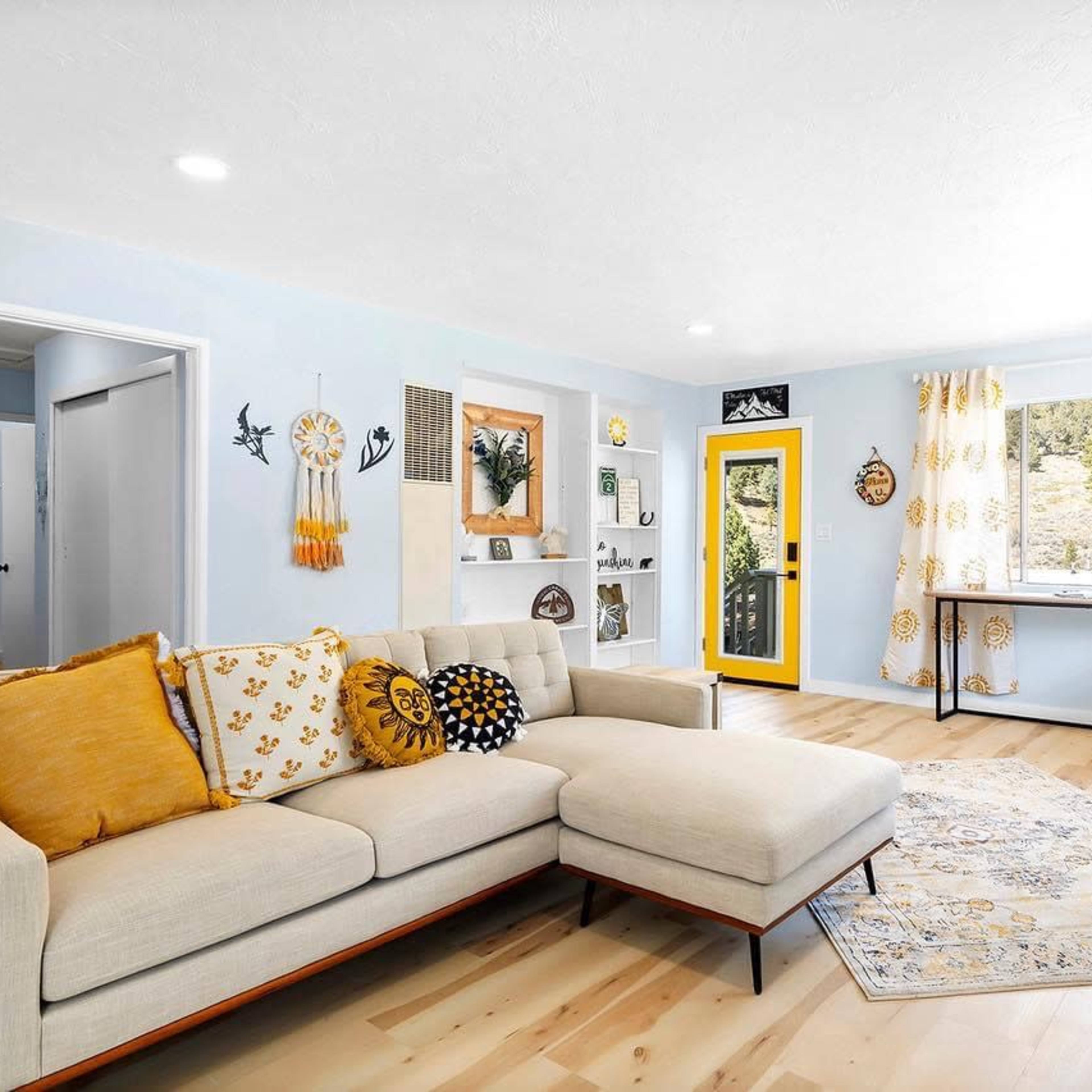 A bright and airy living room features a light-colored sofa with yellow and white accents, a large window with floral curtains, and a cheerful yellow door.