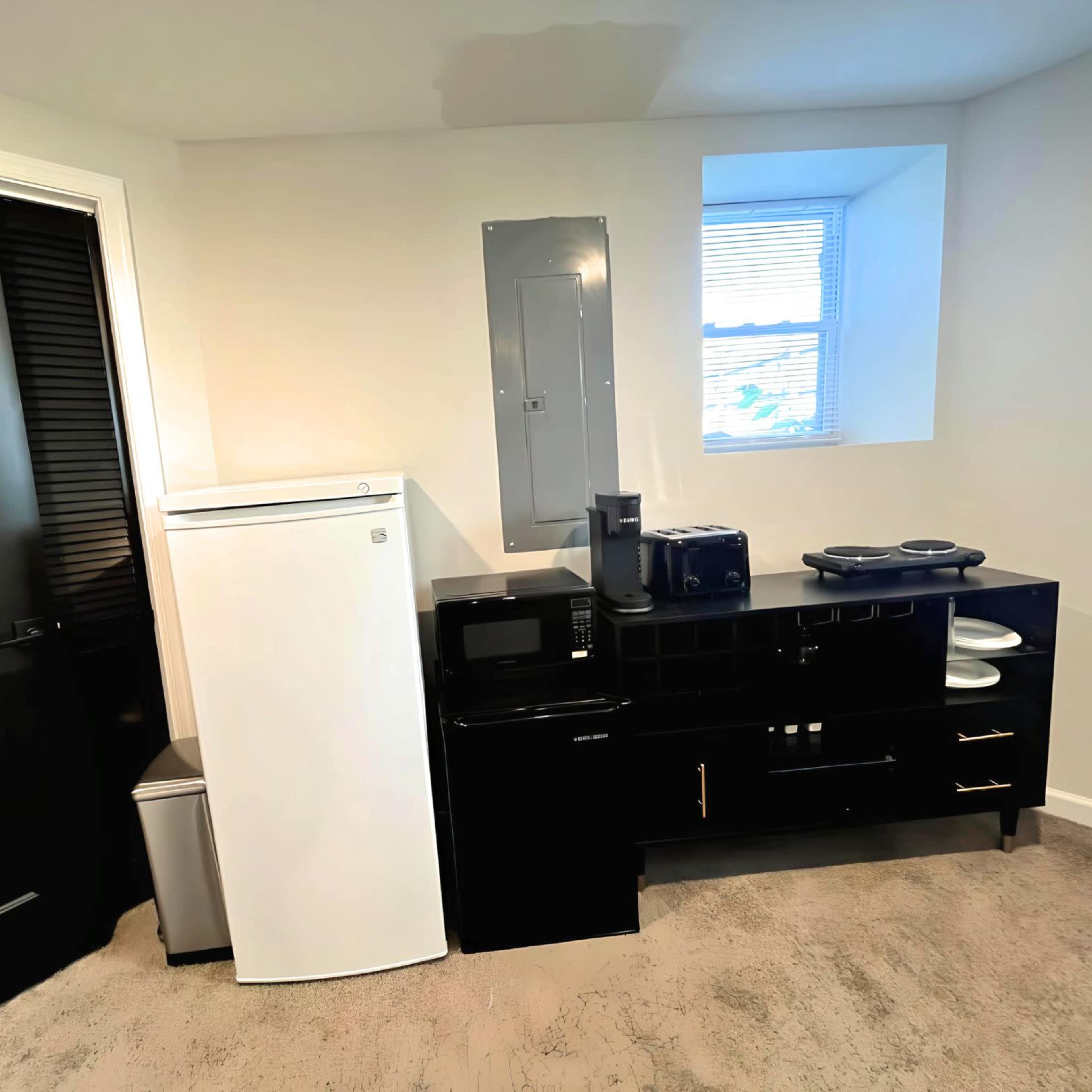 A small kitchenette area featuring a white refrigerator, microwave, and coffee maker against a black cabinet, with a window providing natural light.