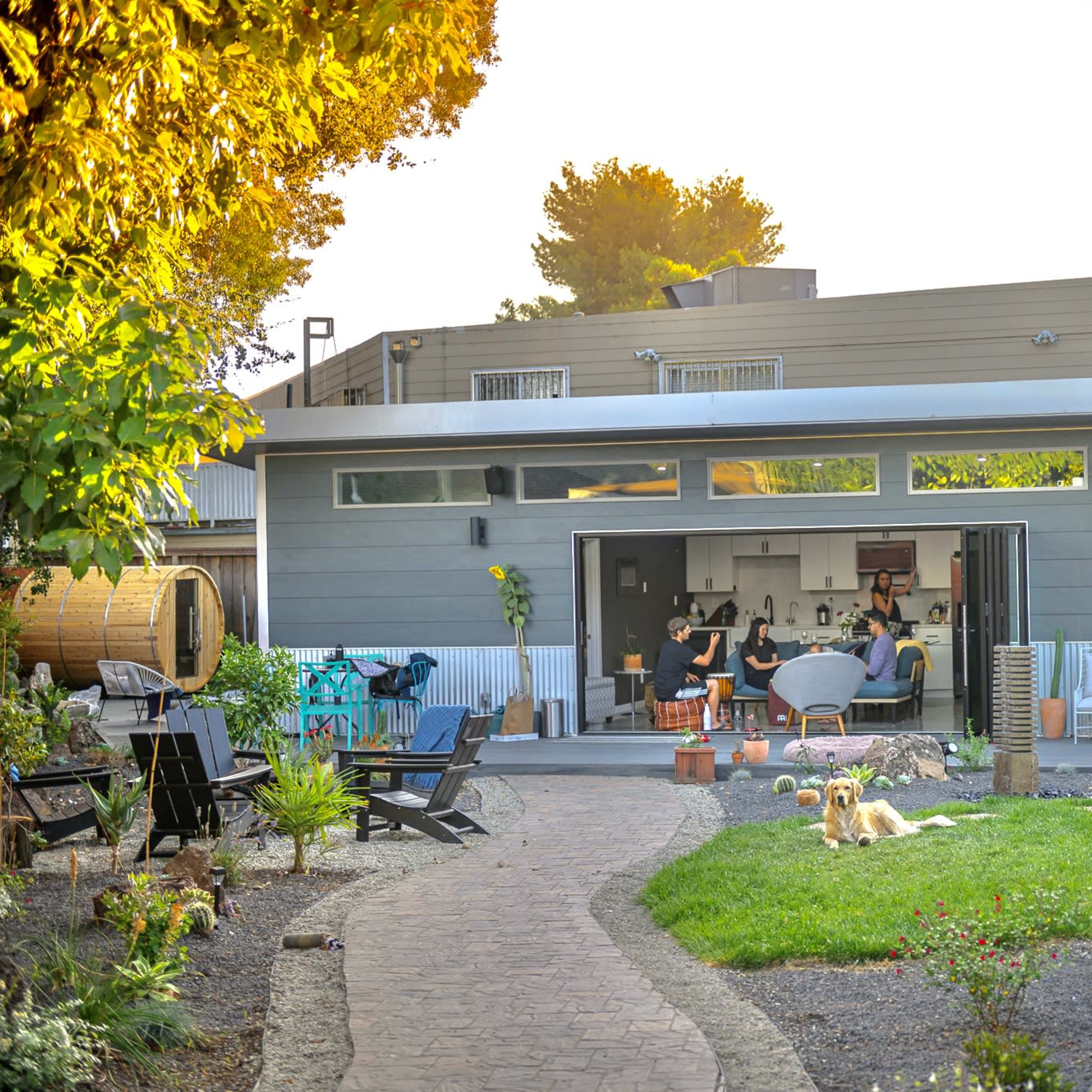 A modern backyard scene features a home with large sliding glass doors opening to a living area, surrounded by greenery and garden decor.