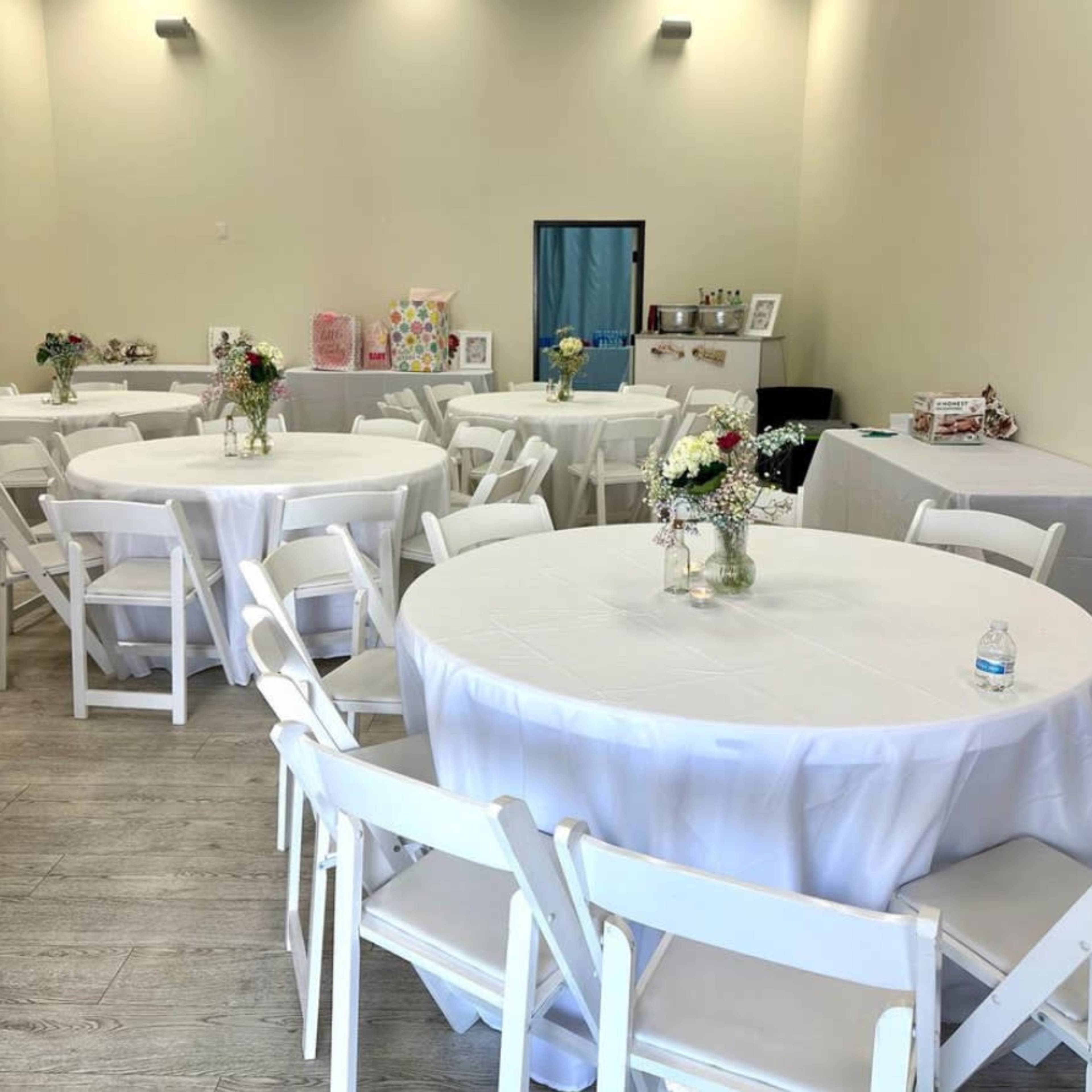 A spacious event room set with several round tables covered in white tablecloths and white chairs arranged neatly around them.