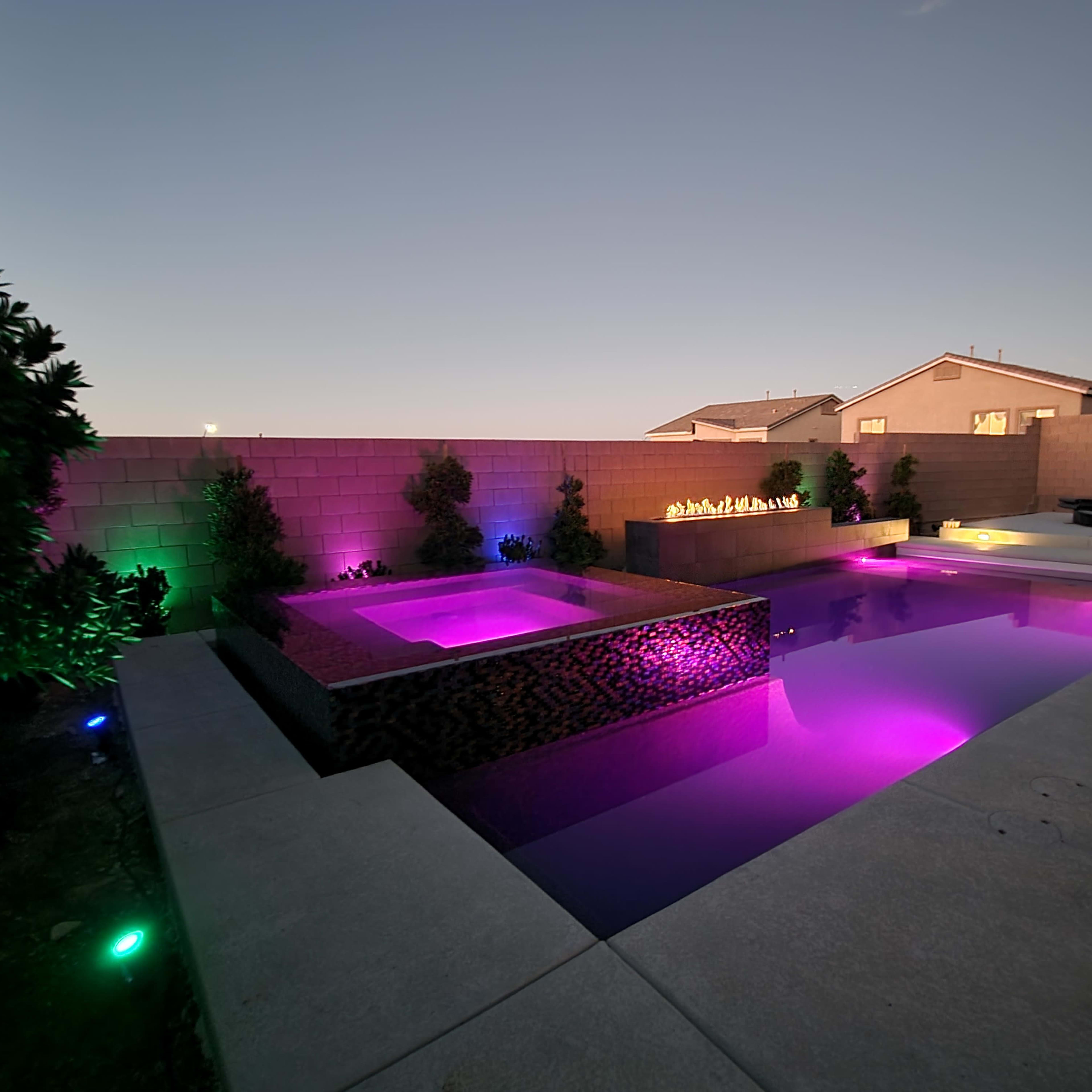 A backyard swimming pool area featuring a lit hot tub and an illuminated pool with colorful lights against a block wall.