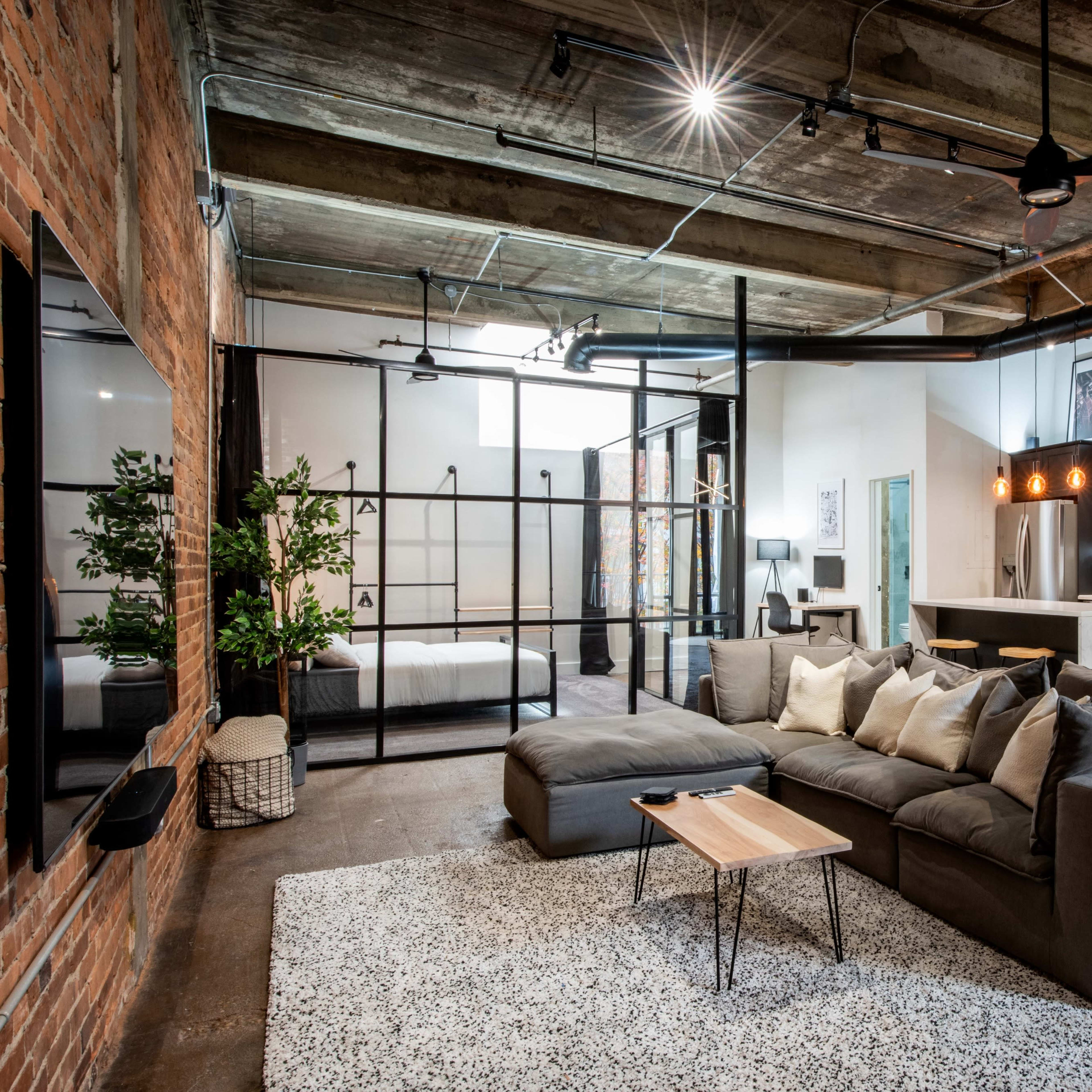 A modern open-concept living space with a sectional sofa, a small wooden coffee table, a kitchen area, and a glass-walled bedroom at the back.