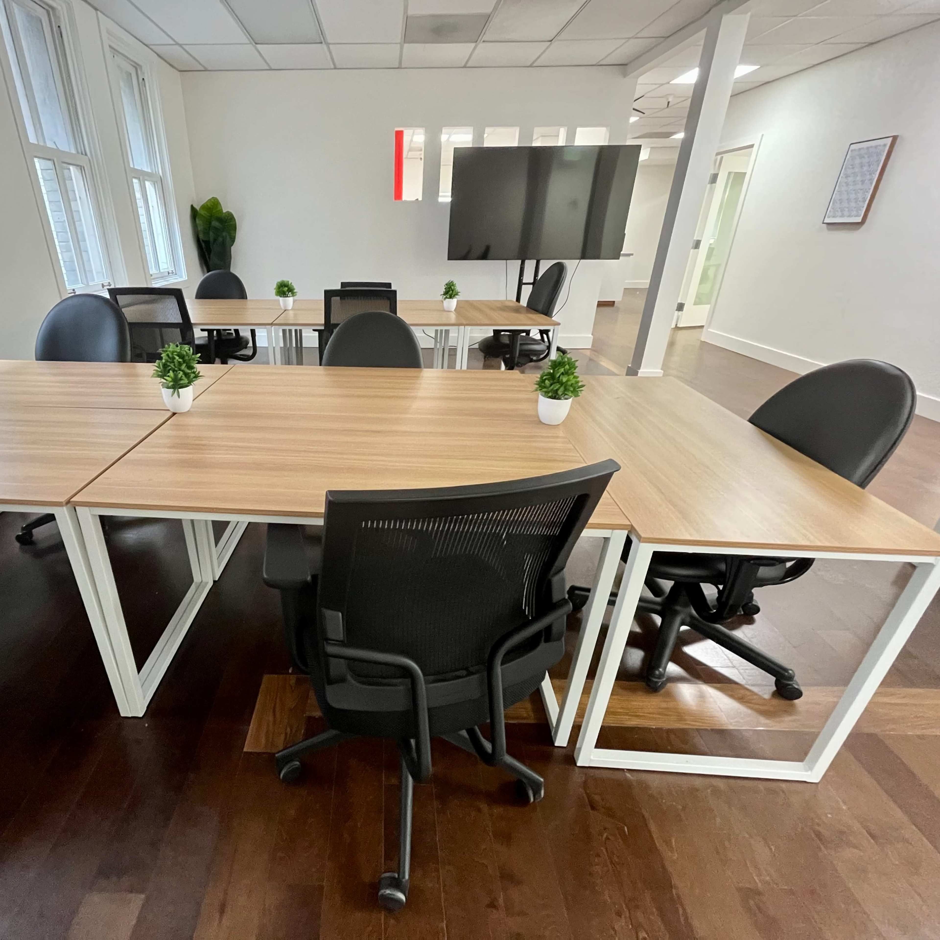 A modern office space features several tables with black chairs and small potted plants, surrounded by large windows and a wall-mounted screen.