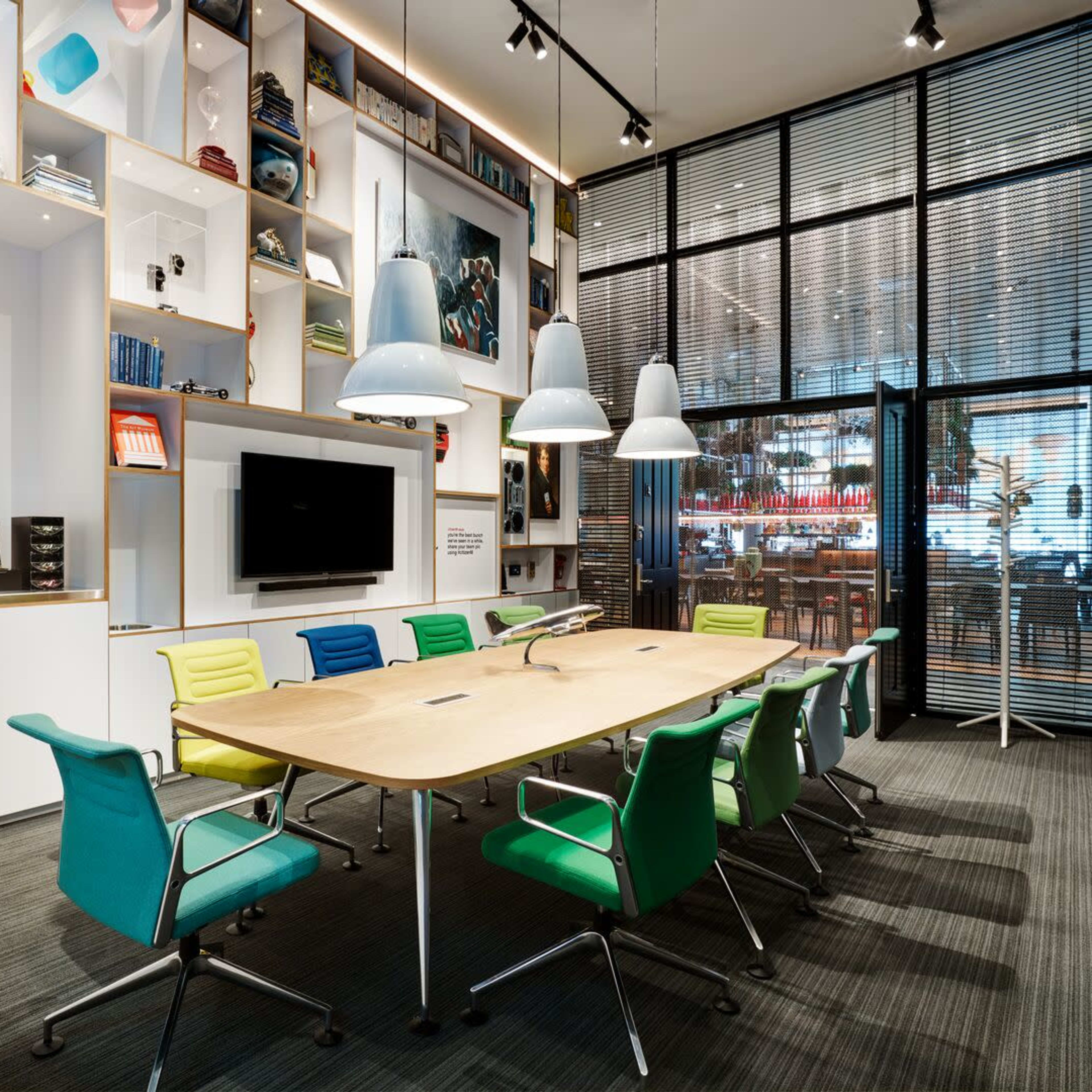 A modern conference room featuring a large wooden table surrounded by colorful chairs, a wall of shelves filled with books and decor, and glass doors leading to an adjacent area.