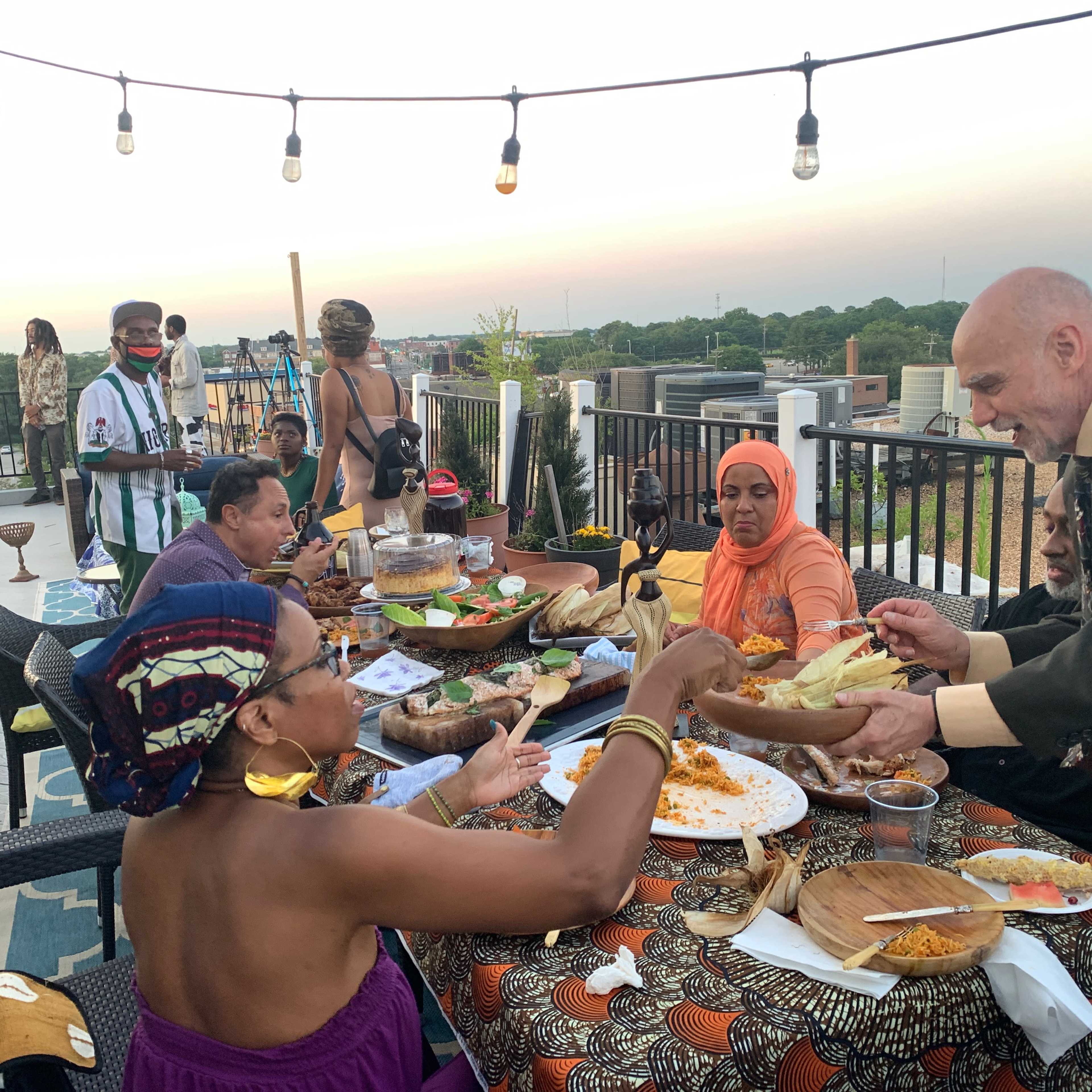 A diverse group of people shares a meal at a rooftop gathering, with food laid out on a table and city views in the background.