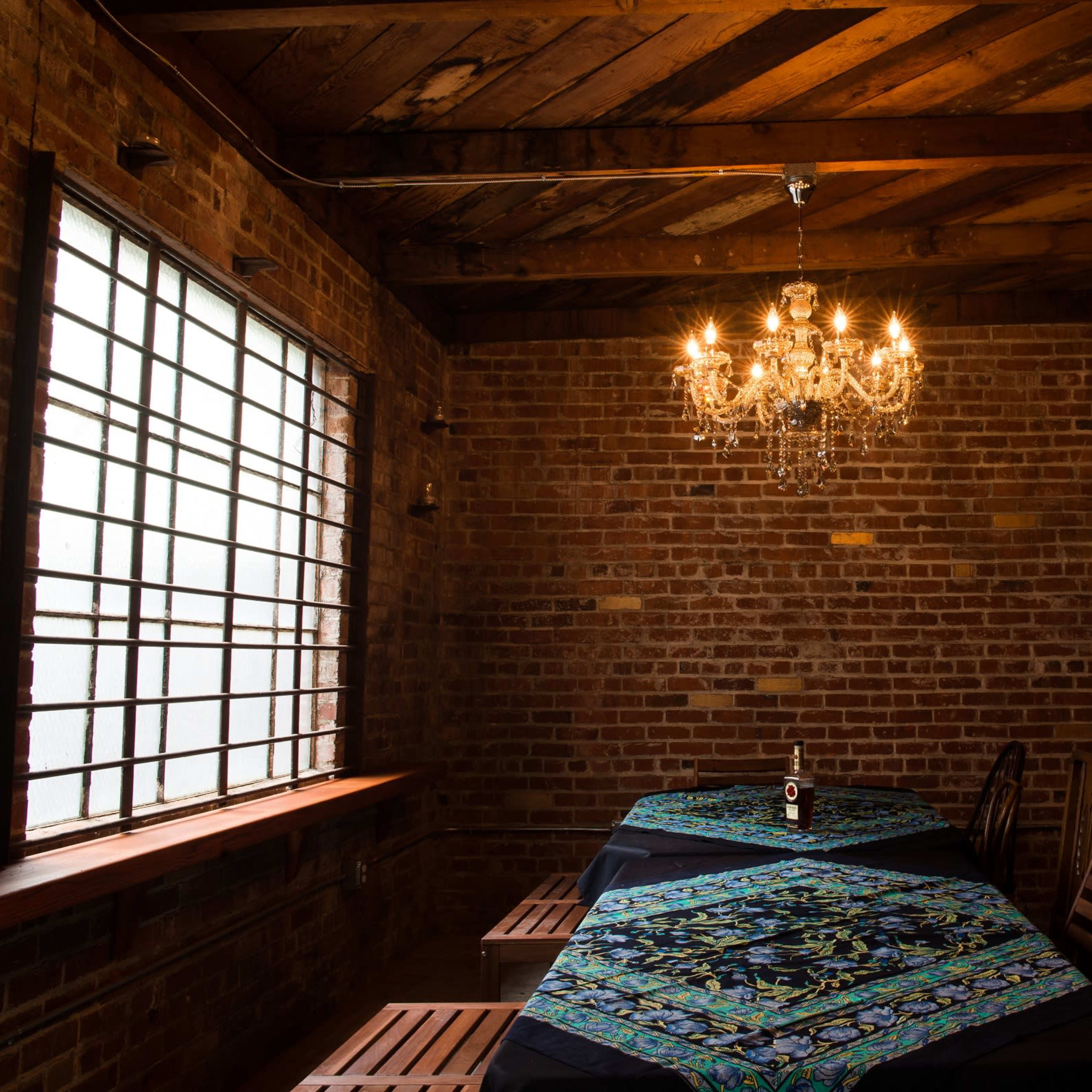 A wooden table is set beneath a chandelier in a brick-walled room with a window and shelves of bottles.