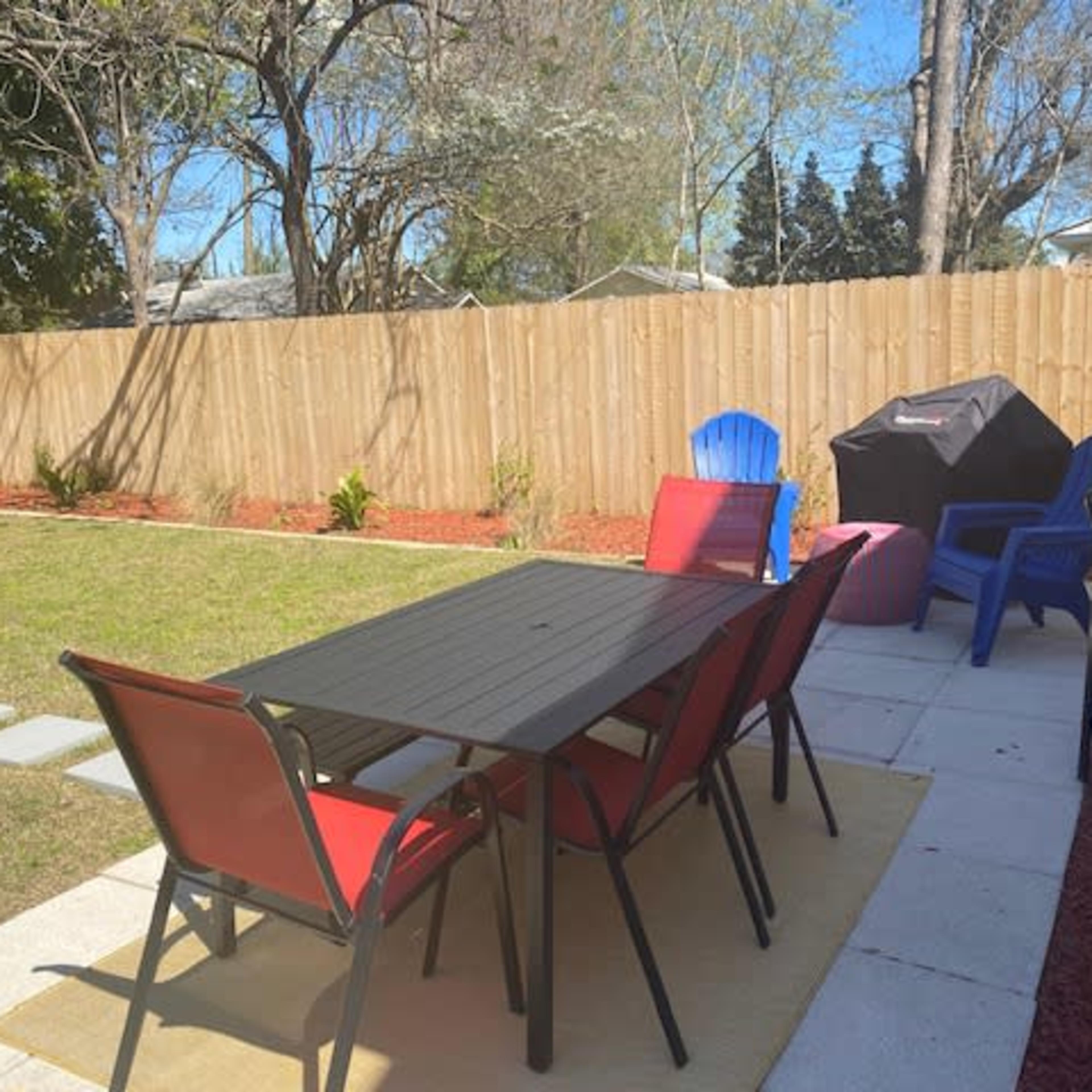 A backyard scene features a dining table surrounded by red chairs, a grill in the corner, and a fenced area with grass and garden beds.