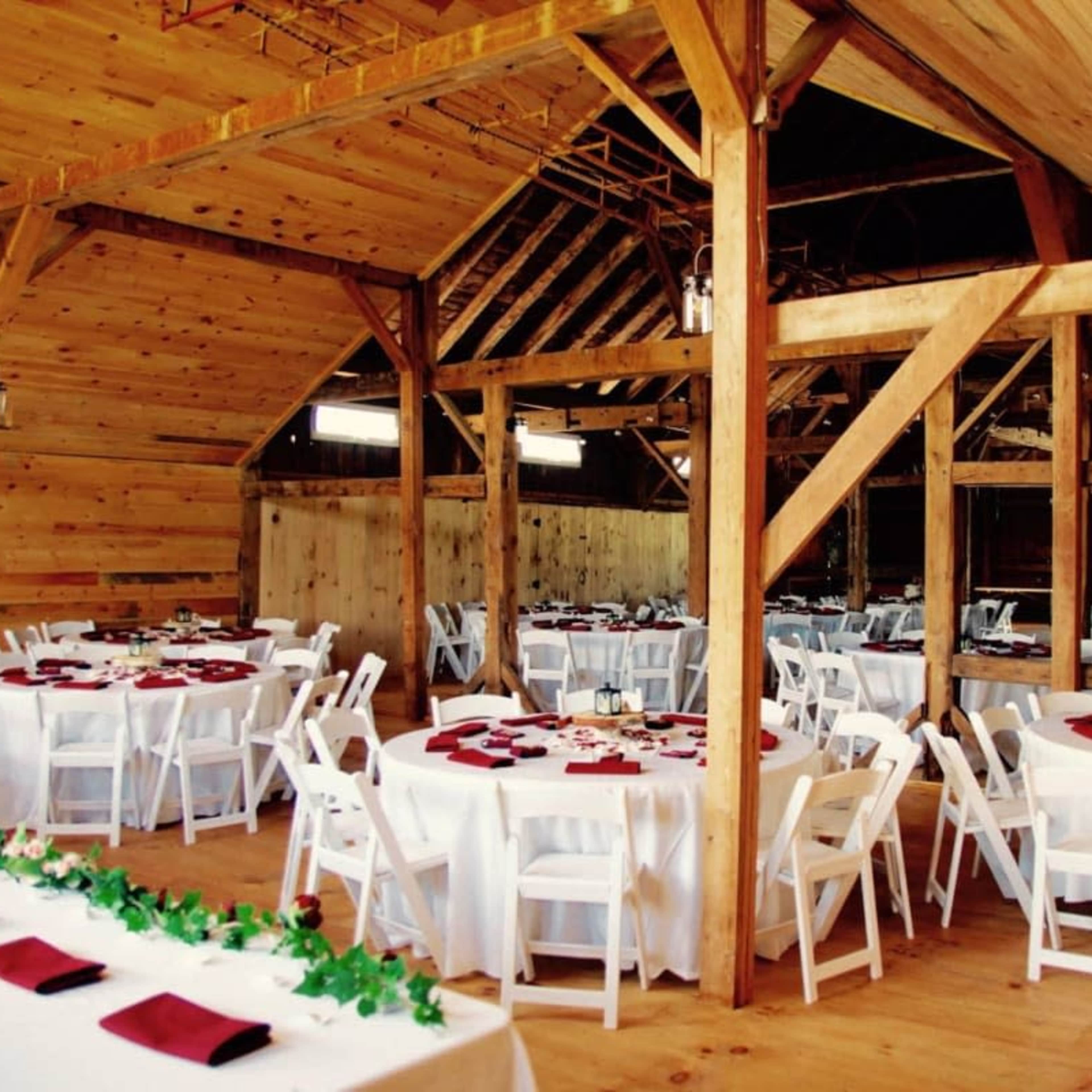 A rustic barn interior set up for a formal dining event, featuring round tables with white tablecloths and maroon napkins arranged around the space.