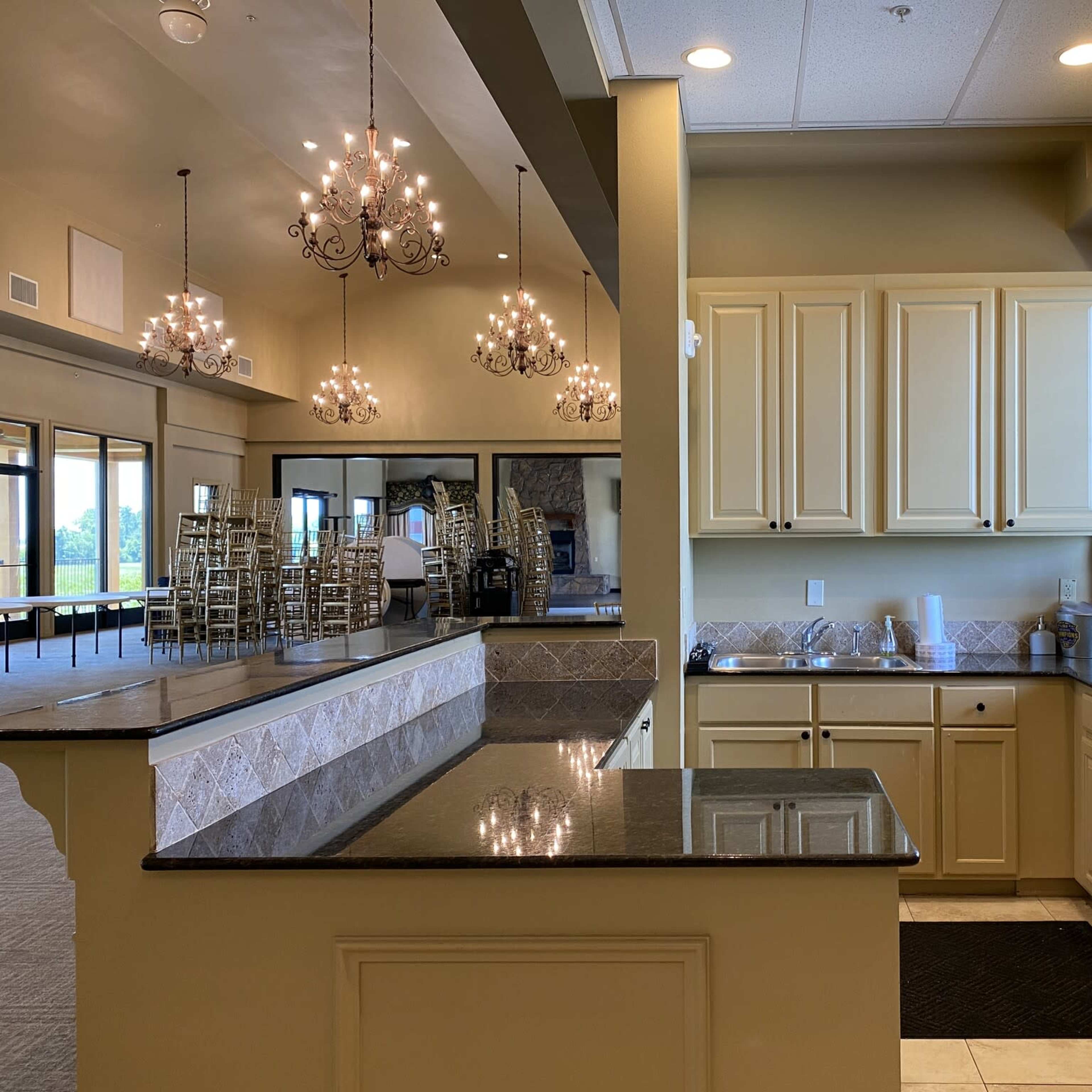 A spacious kitchen area with cream-colored cabinets, a dark countertop, and large windows overlooking a banquet space with chandeliers and stacked chairs.