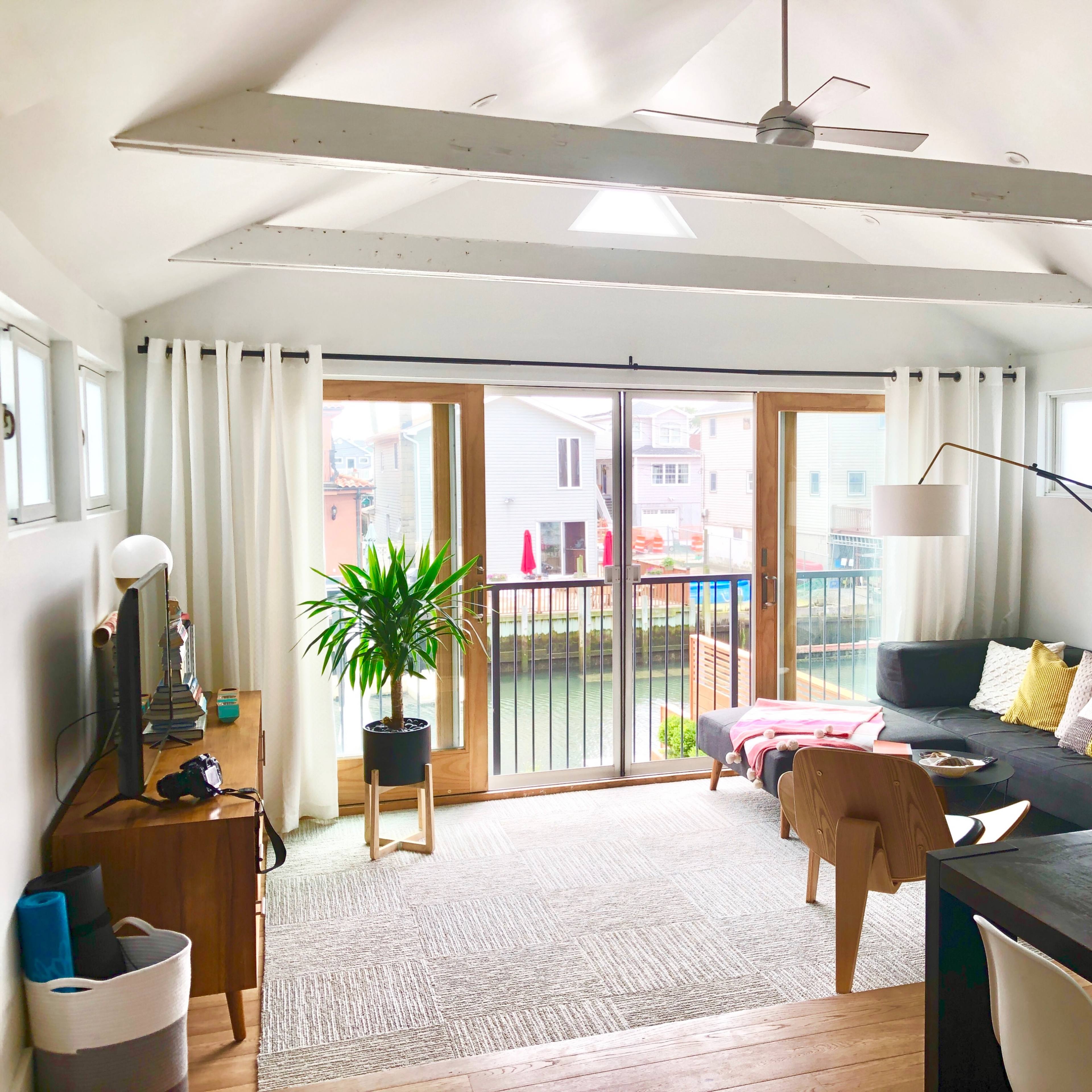 The living room features a sectional sofa, a dining table, and large windows overlooking a deck with a view of nearby houses.