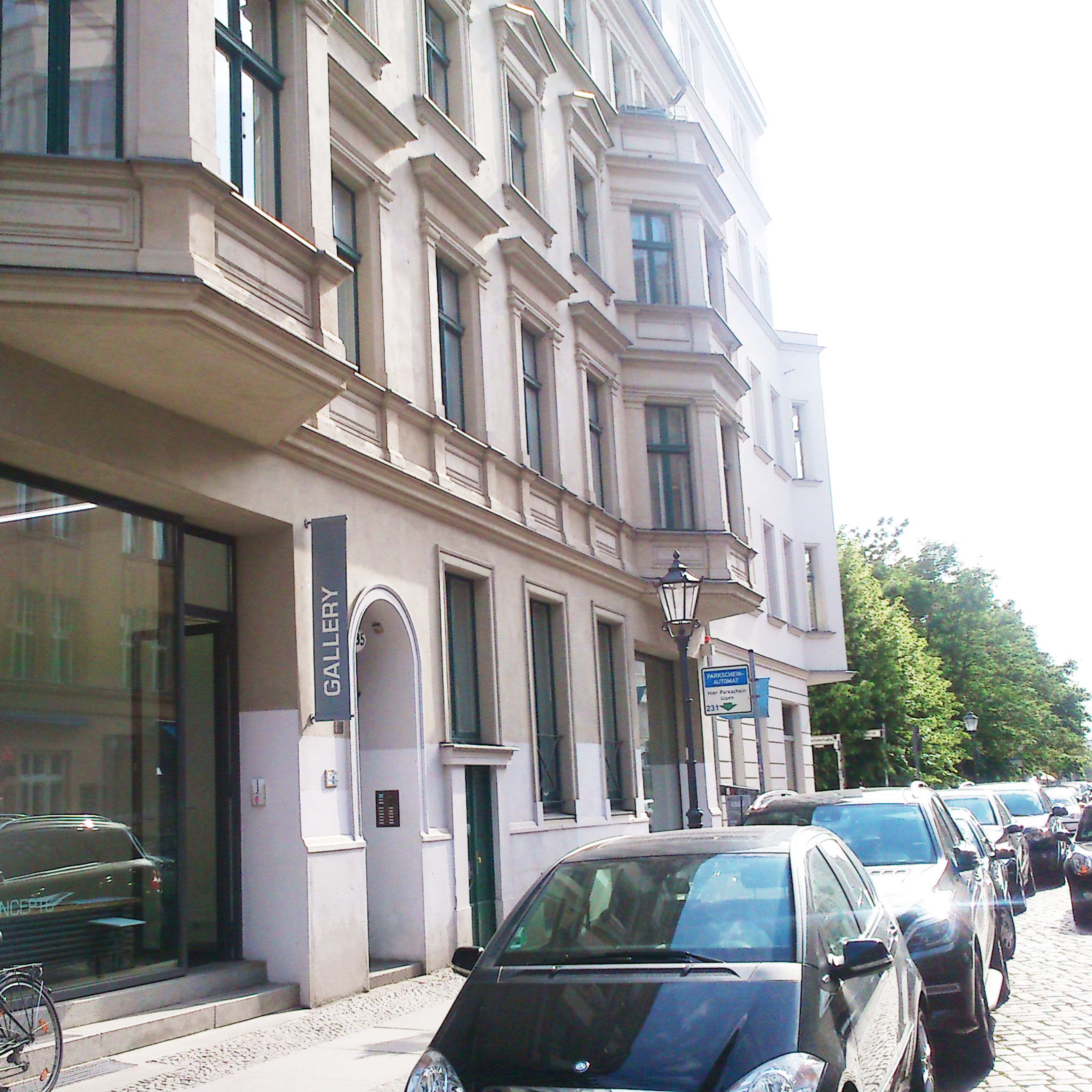 A street view of a multi-story building with a large glass window displaying a sign that reads "GALLERY," surrounded by parked cars and trees.