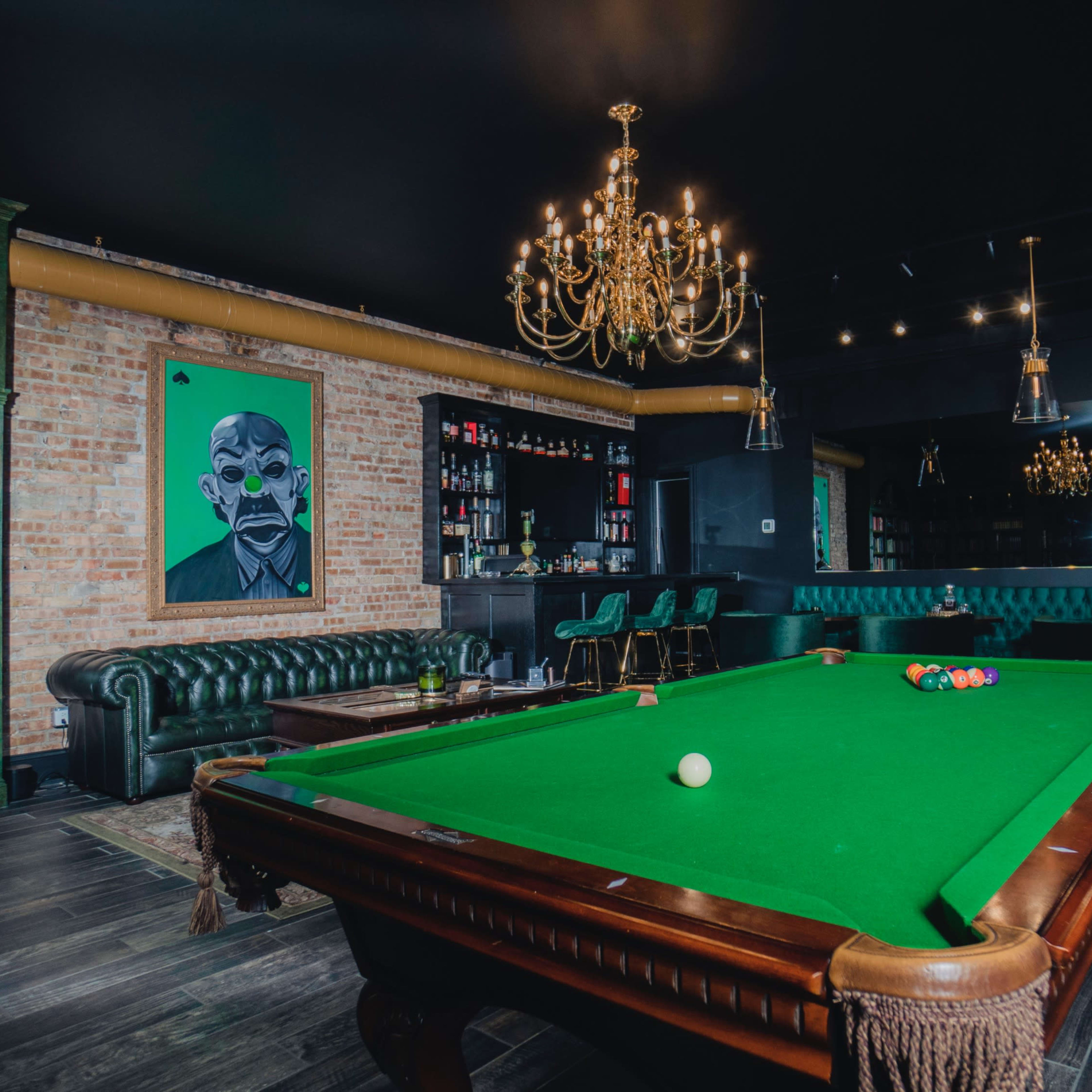 A spacious game room features a green billiards table, a classic leather sofa, shelves lined with books, and a striking portrait on the wall.