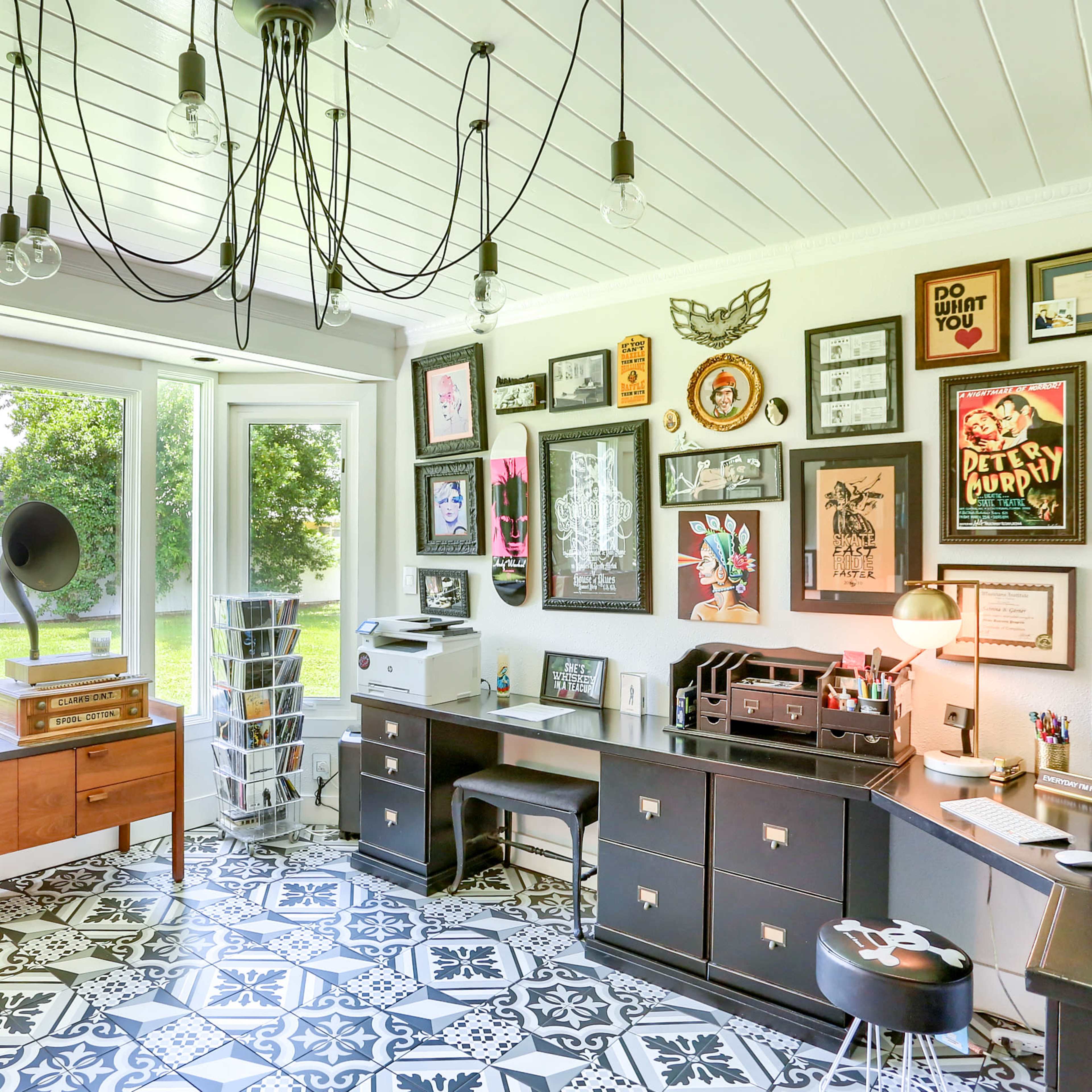 A brightly lit home office features two desks, a large wall of framed artwork, and a patterned tile floor, with windows overlooking a green yard.