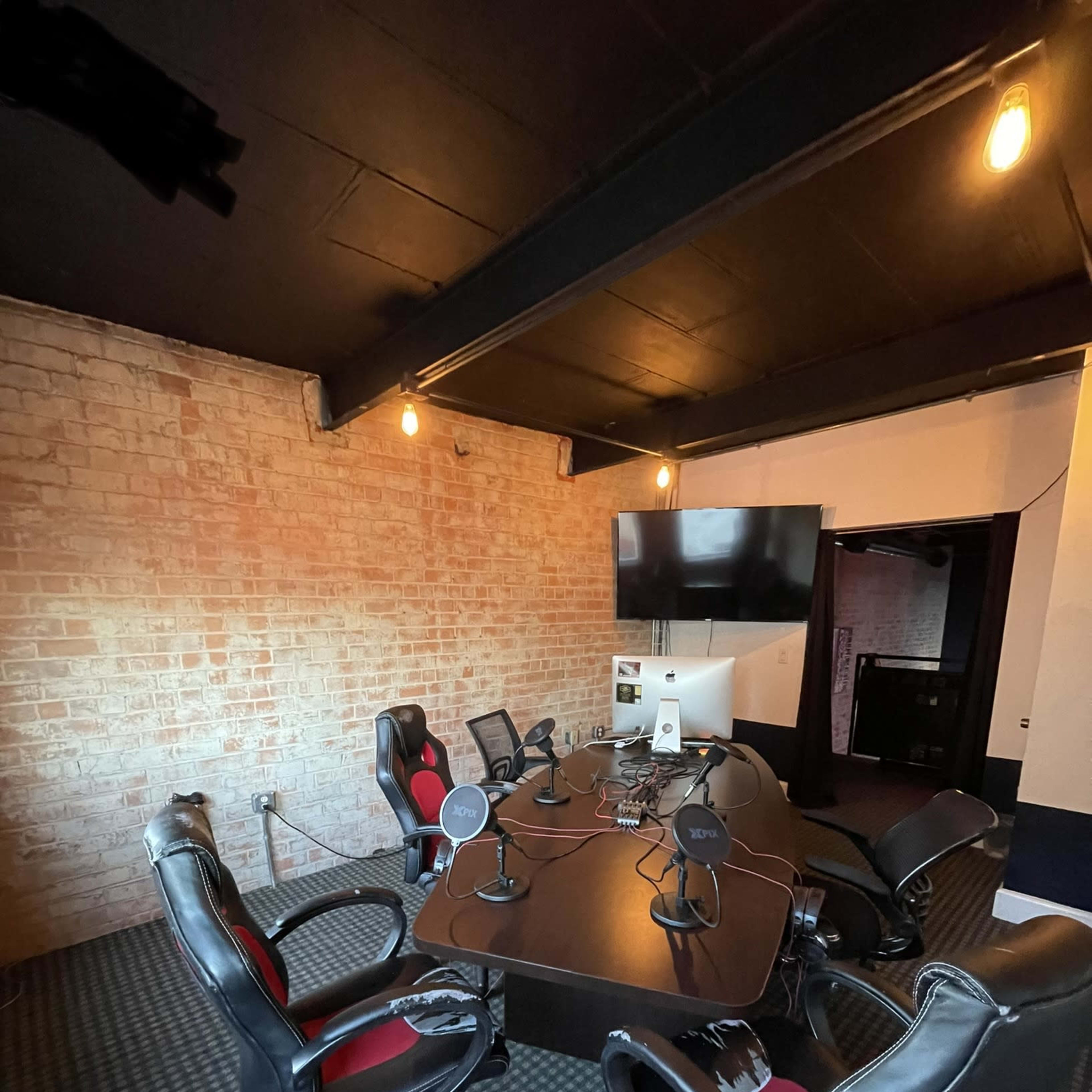 An office room with a large conference table surrounded by several black and red chairs, equipped with microphones and a computer, set against a brick wall.
