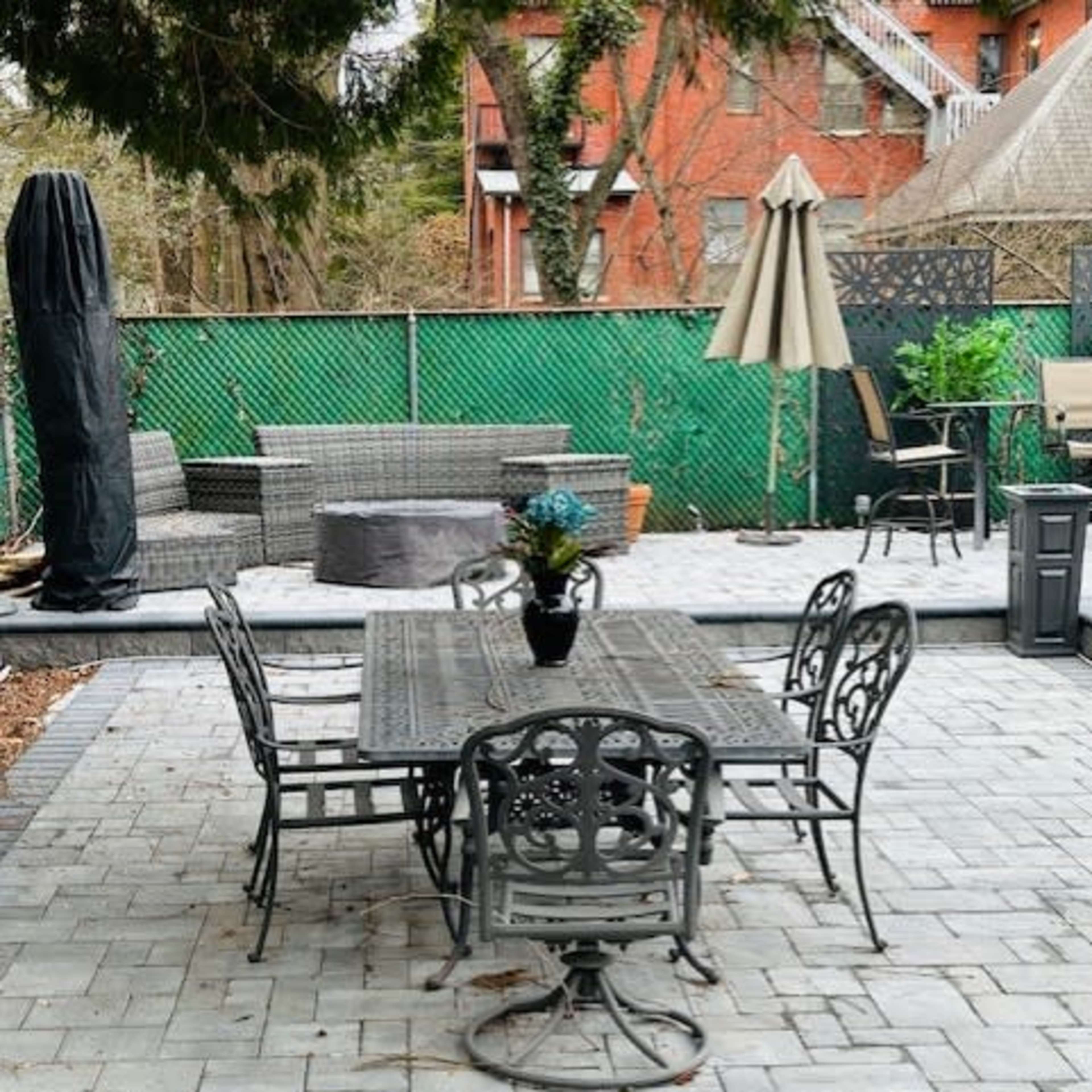 A patio area features a metal dining table surrounded by five chairs, with a large umbrella and various seating arrangements in the background.