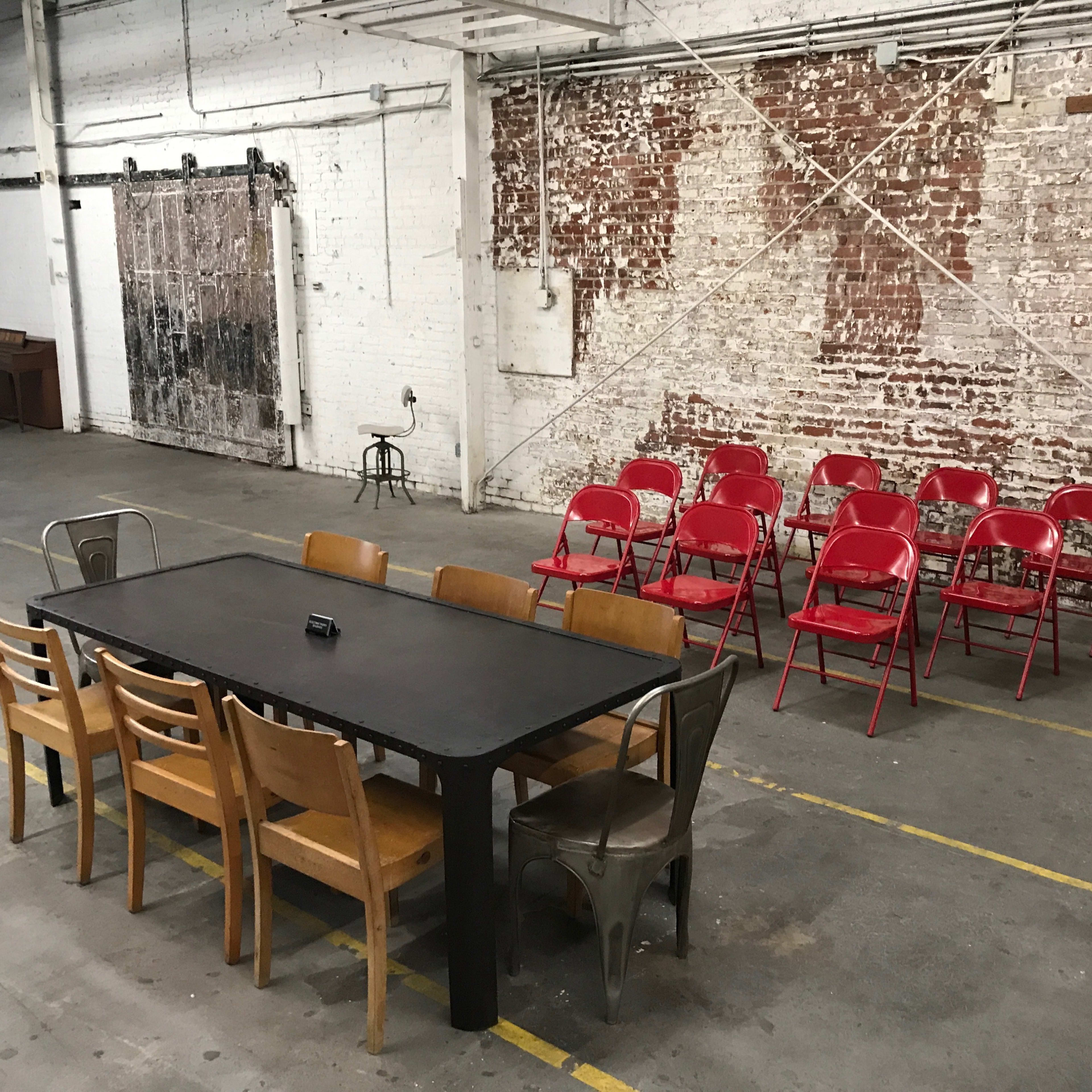 A long black table with wooden chairs is positioned in front of a row of red folding chairs against a brick wall in an industrial space.