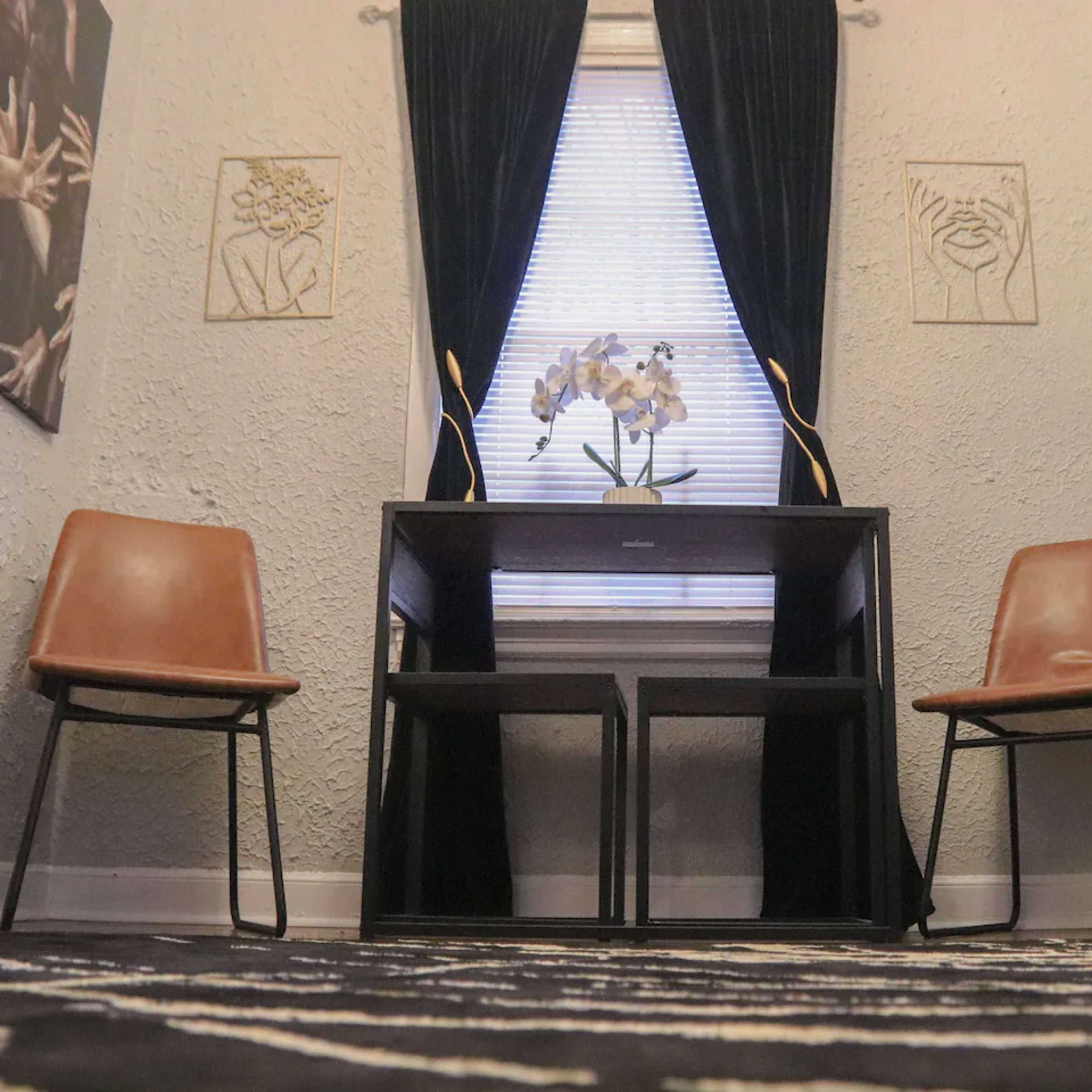 A small room with a desk and two chairs facing each other, adorned with wall art and a window draped with black curtains.