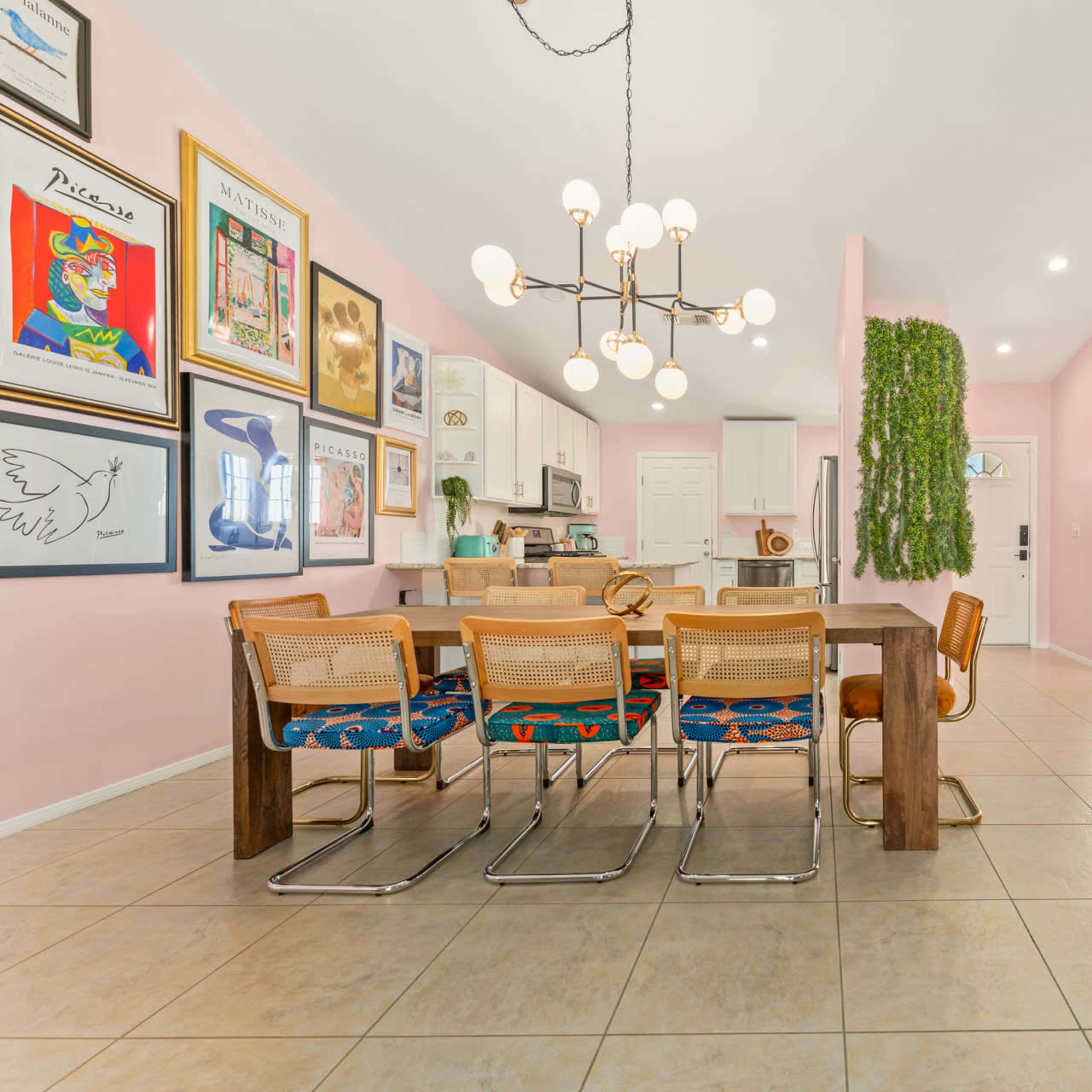 A dining area with a large wooden table surrounded by chairs, colorful artwork on the walls, and a pink color scheme.