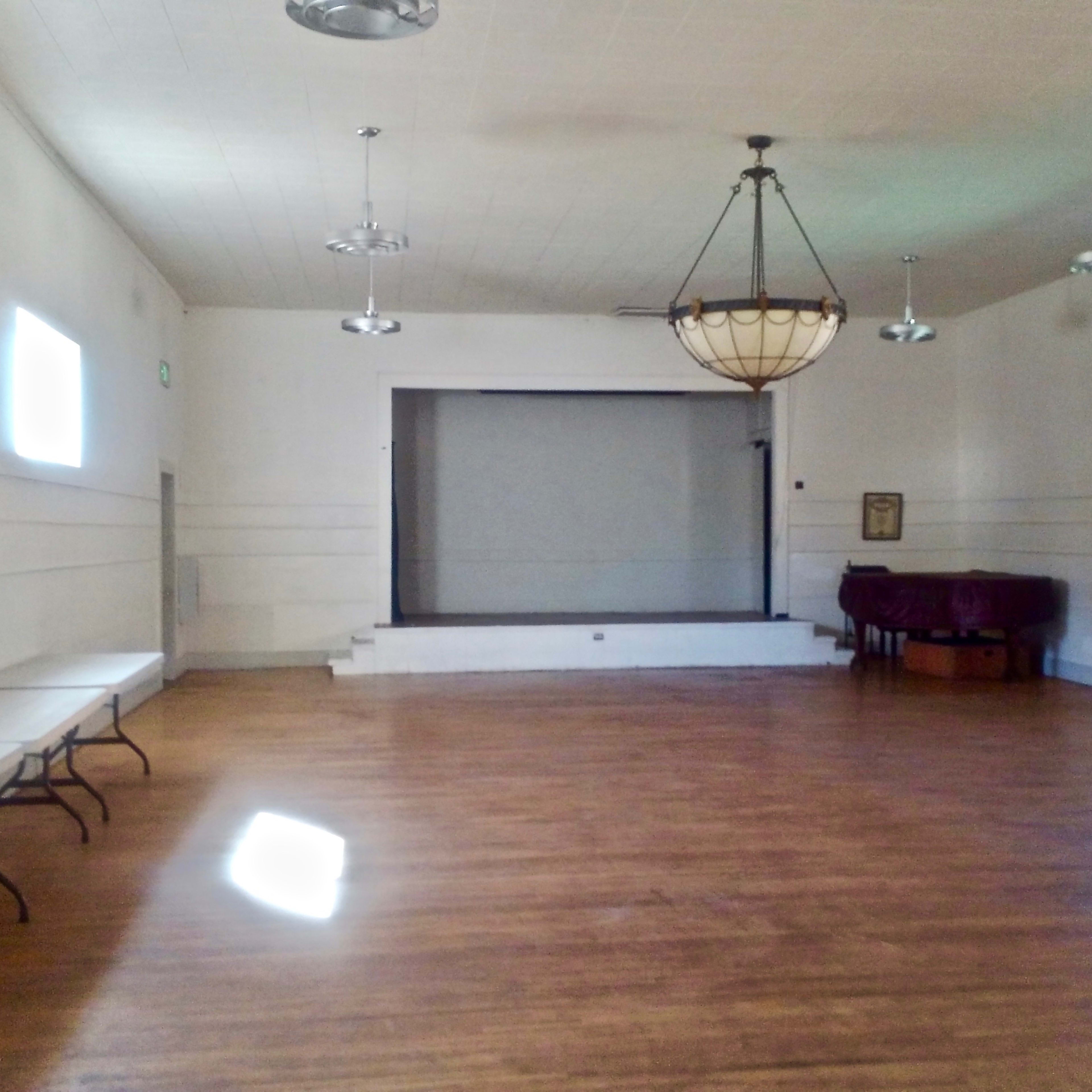 A spacious, empty hall with wooden floors, a chandelier, and folding tables along one side.