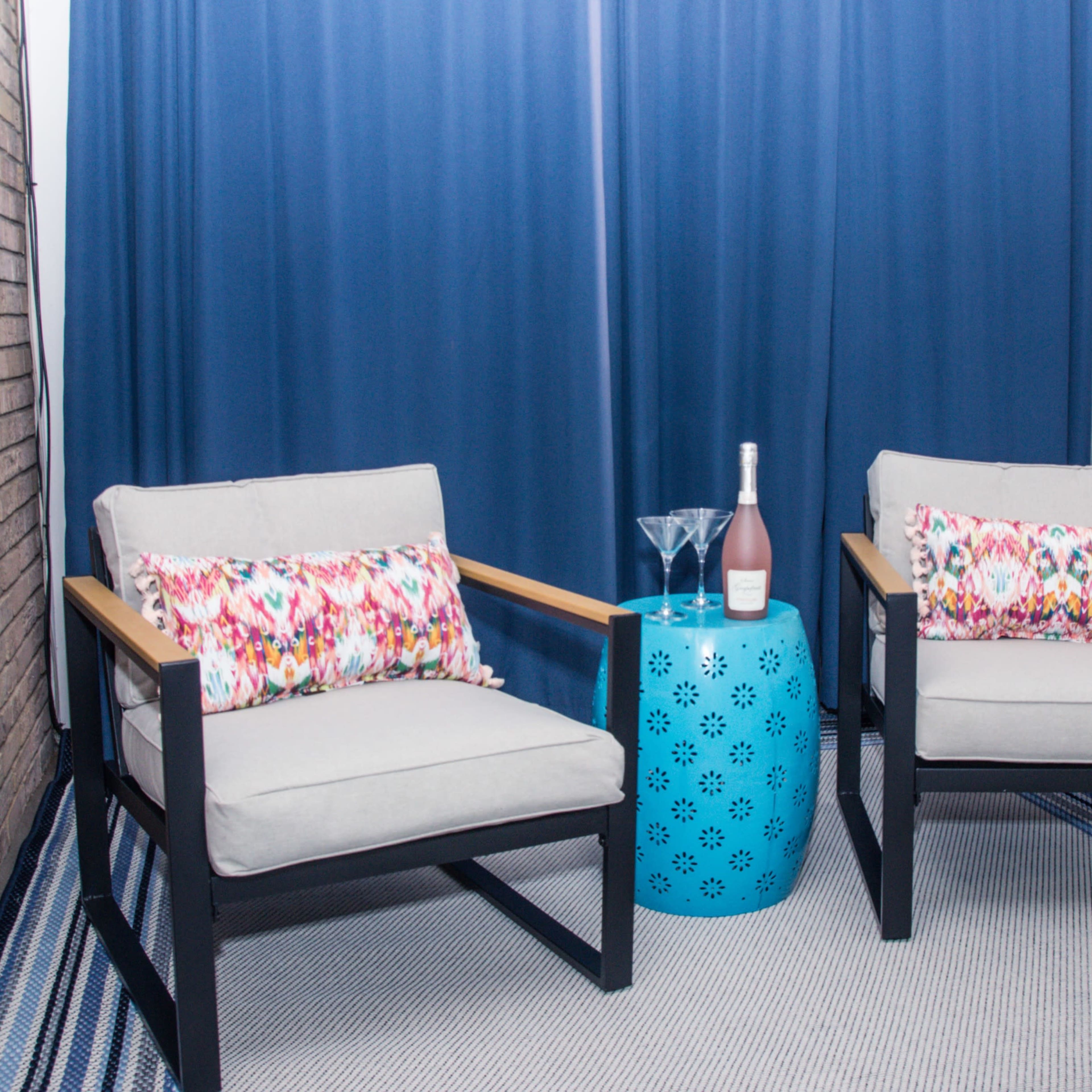 A cozy seating area features two gray armchairs with colorful cushions beside a blue decorative table, set against a backdrop of blue curtains and a brick wall.
