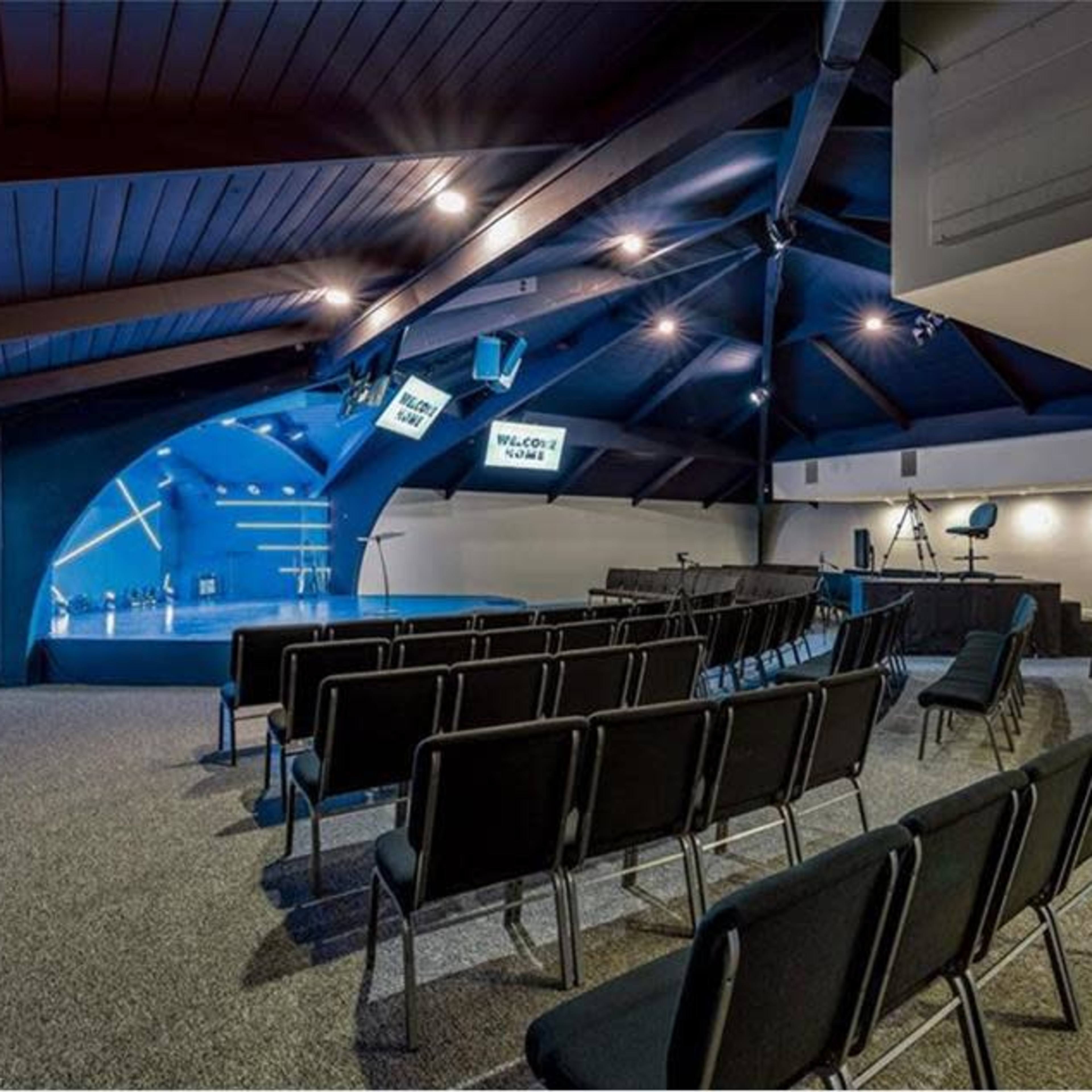 A spacious event room with black chairs arranged in rows facing a stage, illuminated by overhead lights and featuring a large blue wall.