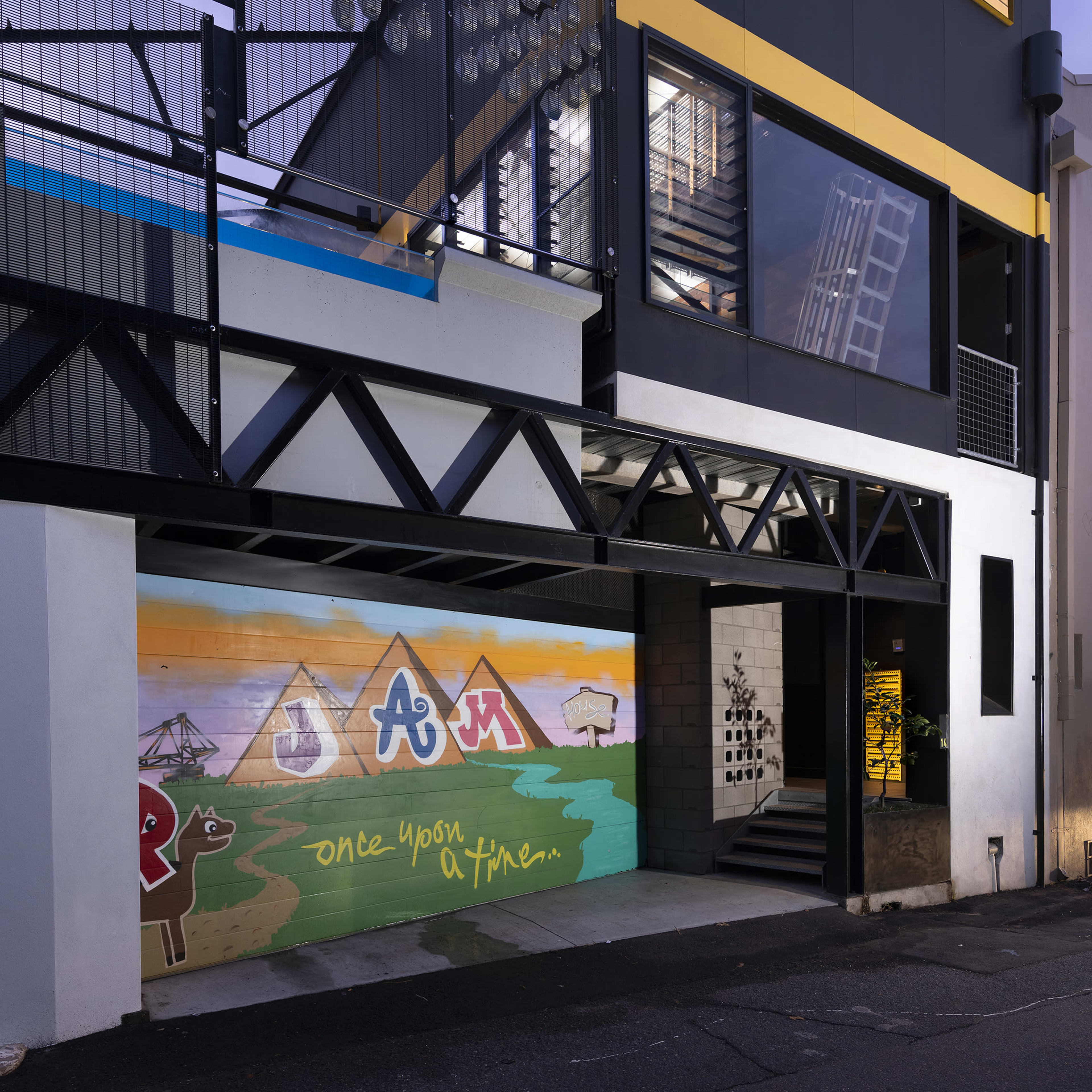 A modern building with a vibrant mural featuring mountains and the word "JAM" on one wall, alongside an industrial-style structure with large windows and metal balconies.