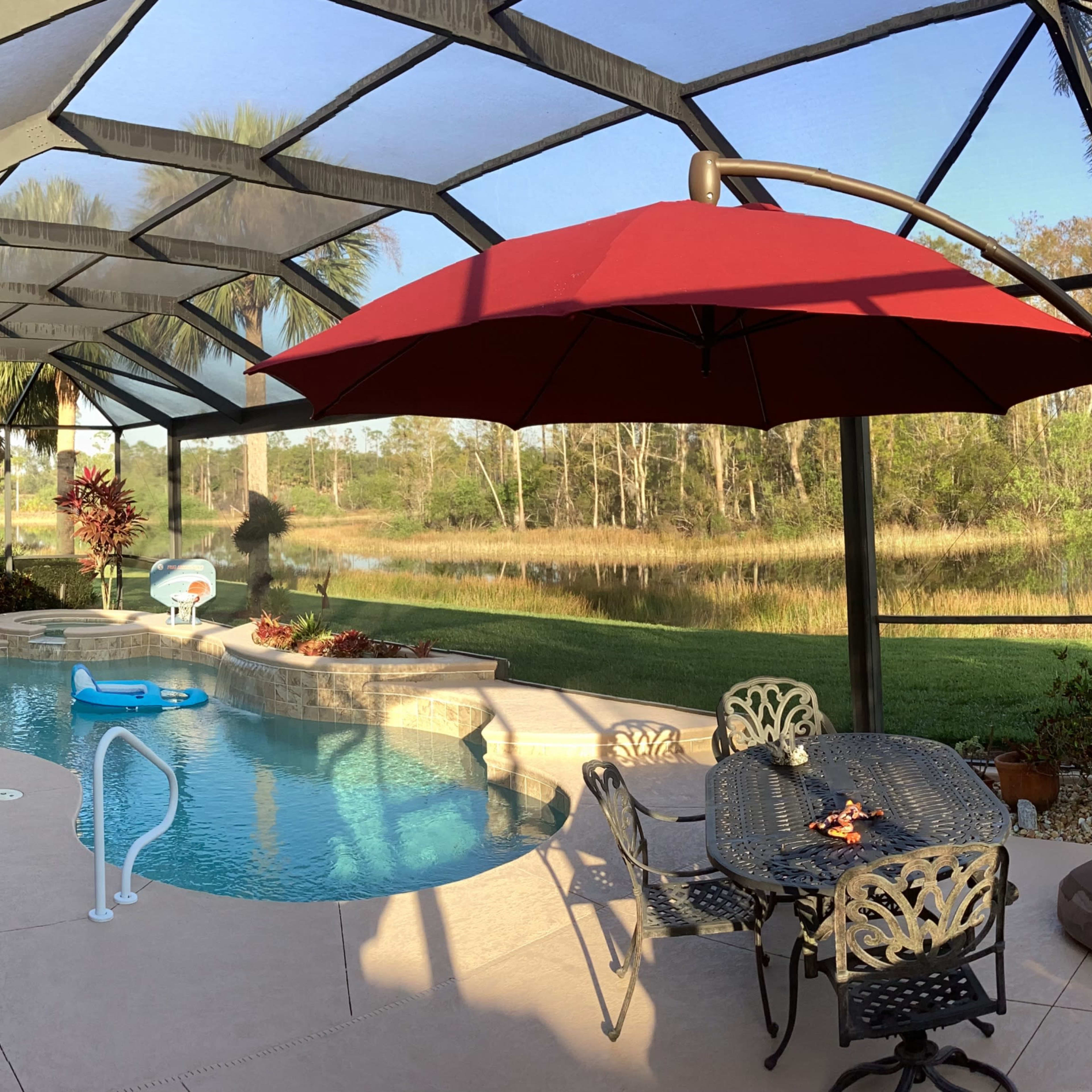 A screened pool area features a swimming pool, a table with chairs, and red umbrellas overlooking a grassy area and water in the background.