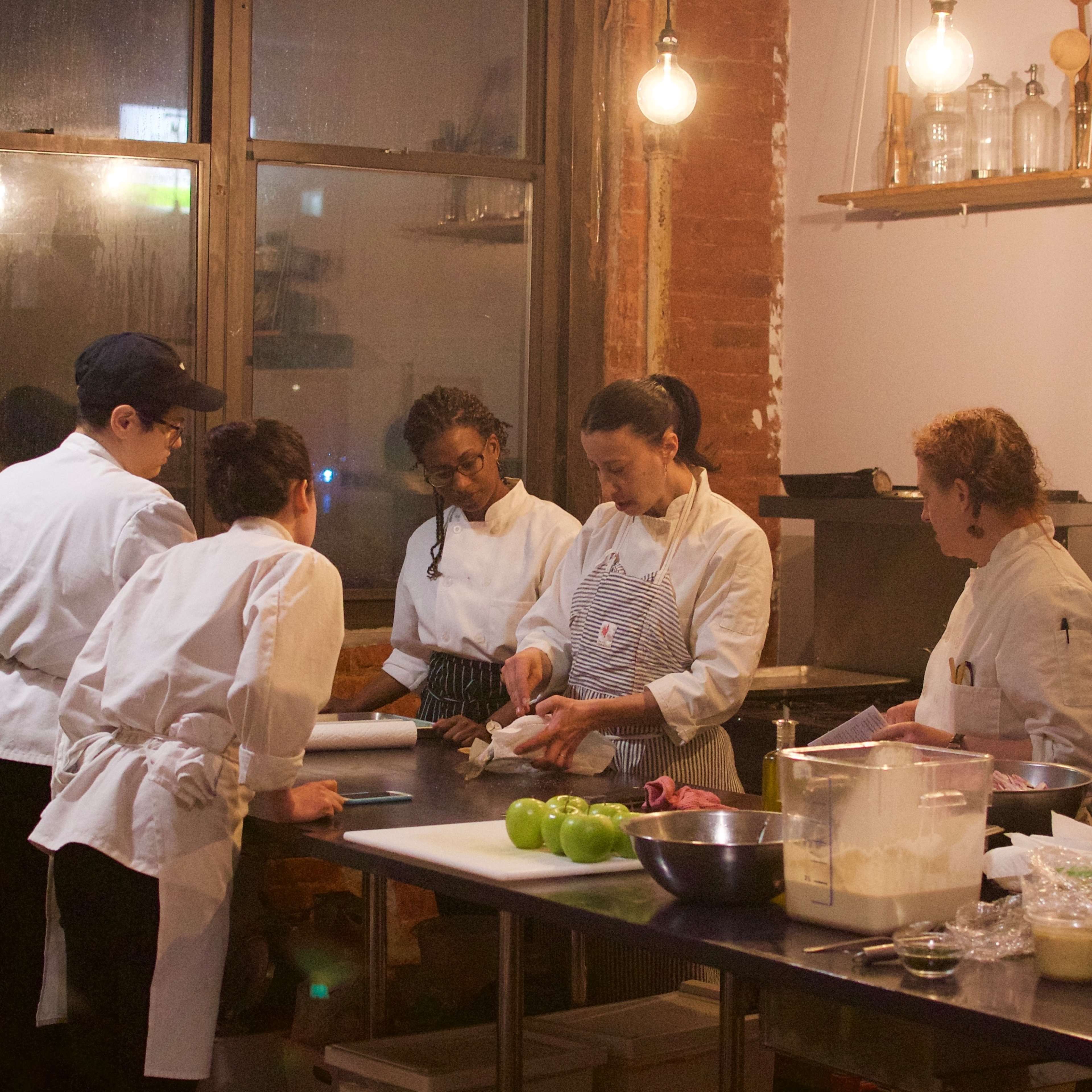 A group of five chefs prepares food in a restaurant kitchen, surrounded by various kitchen equipment and ingredients.