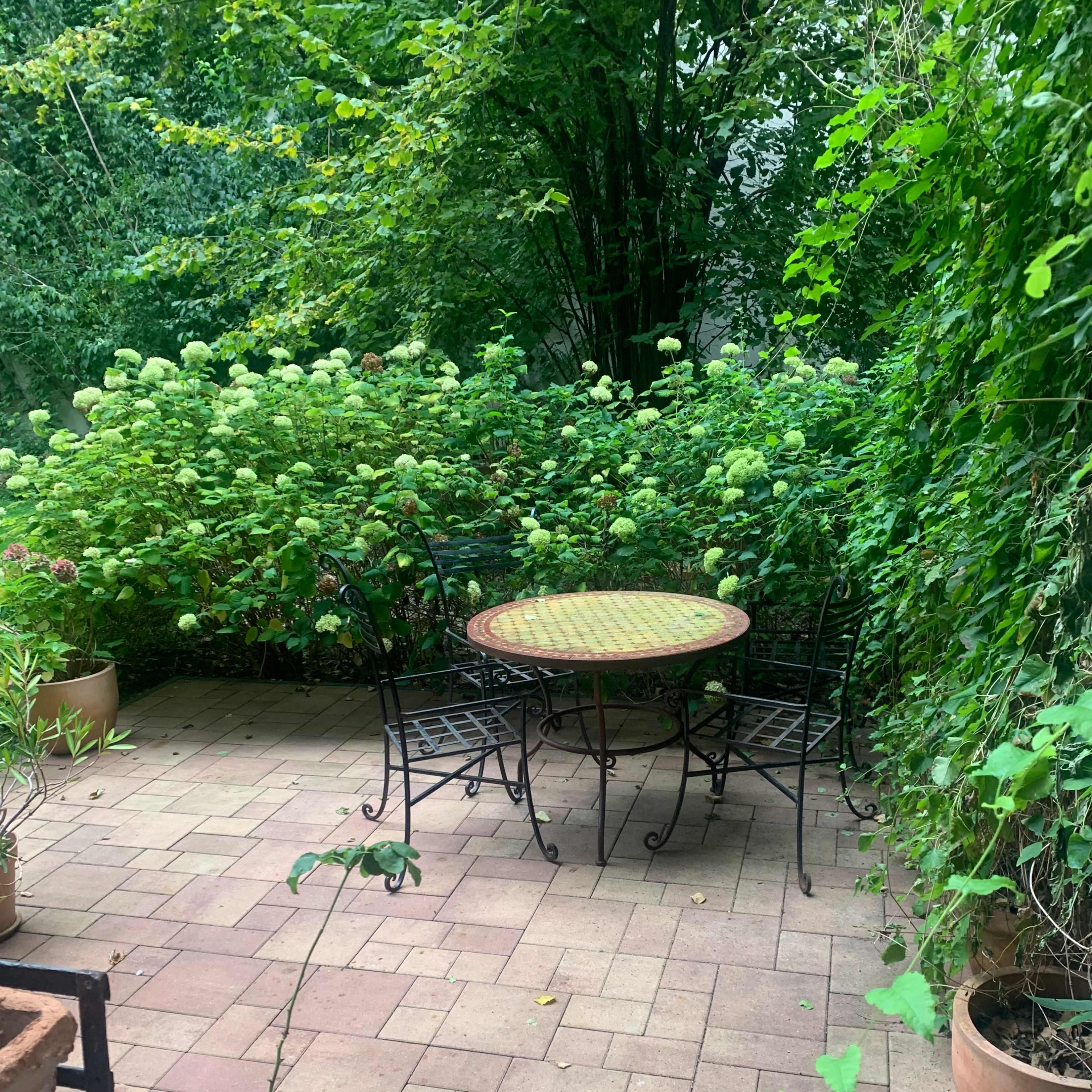 A wrought-iron table and chairs are positioned on a brick patio surrounded by lush green foliage and potted plants.
