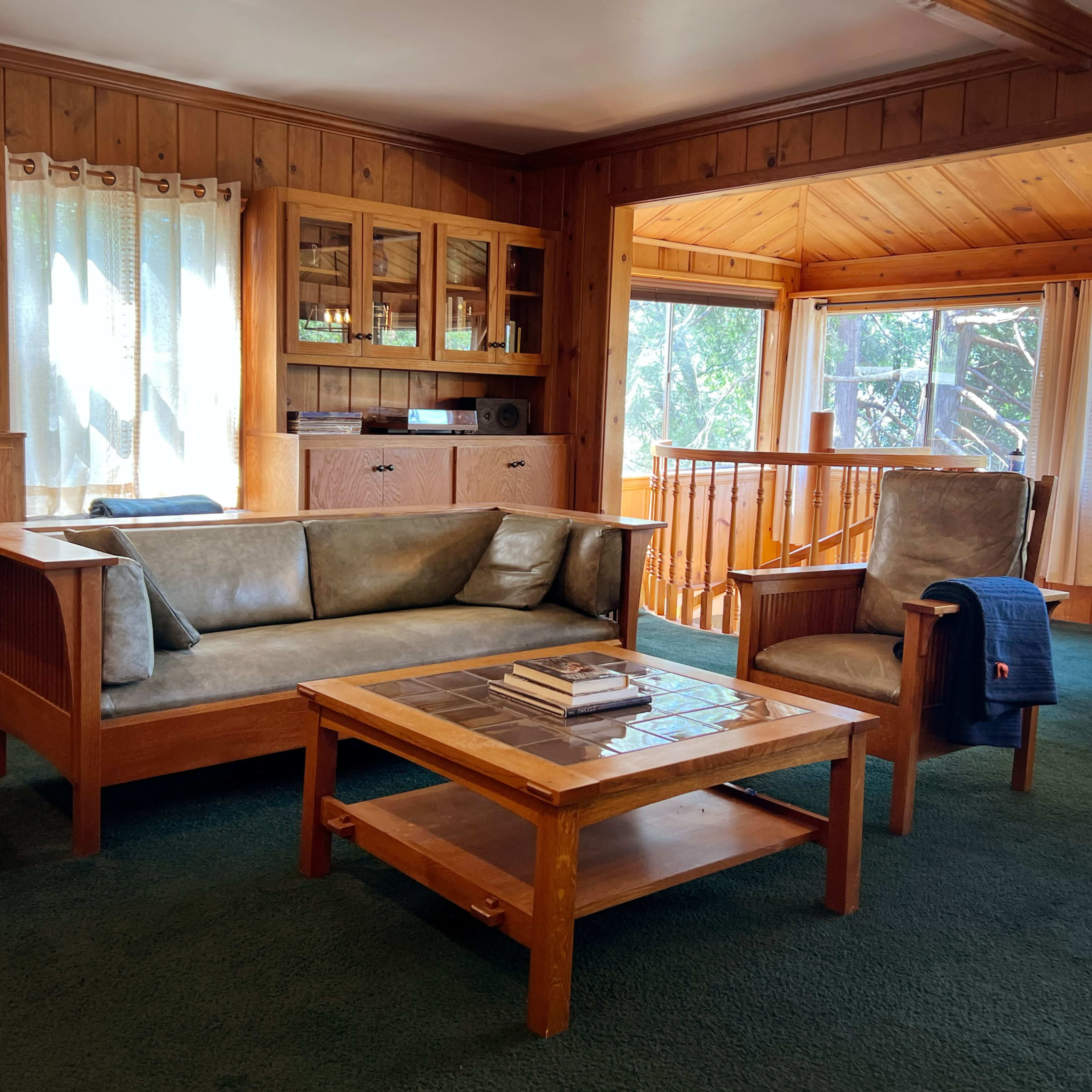 A cozy living room with wooden furniture, a sofa, a coffee table, and large windows letting in natural light.