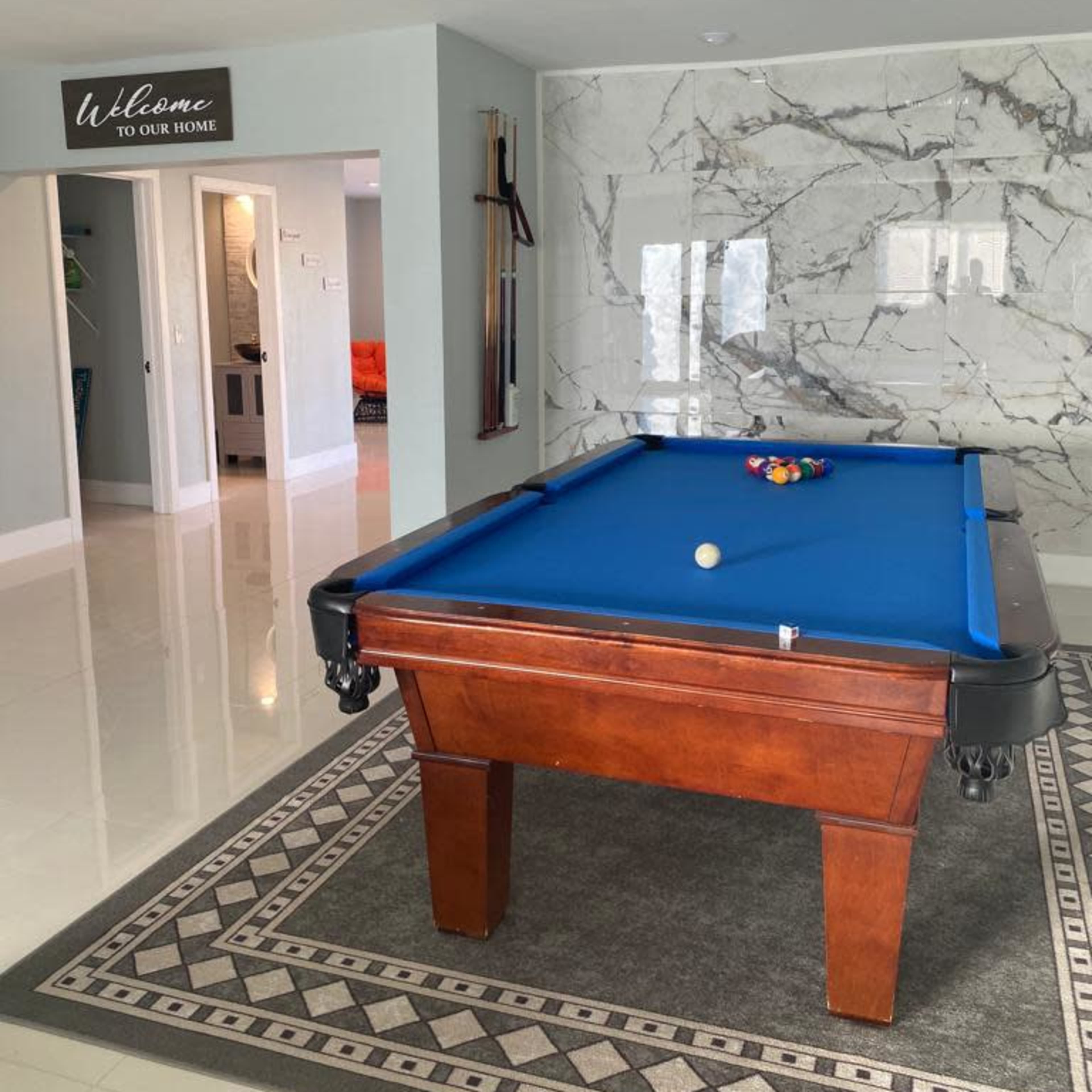 A brightly lit room featuring a blue pool table on a patterned rug, with a marble wall and an entrance leading to another part of the house.