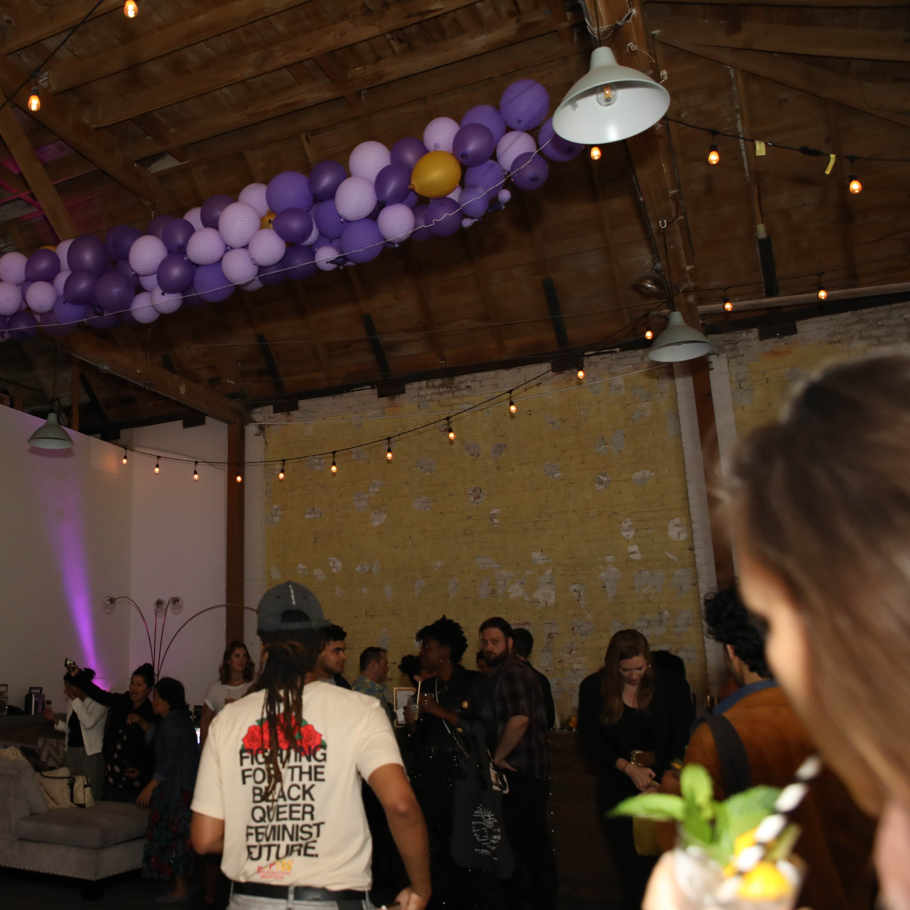 A gathering takes place in a rustic room decorated with purple balloons, where people socialize amidst warm lighting and wooden beams.