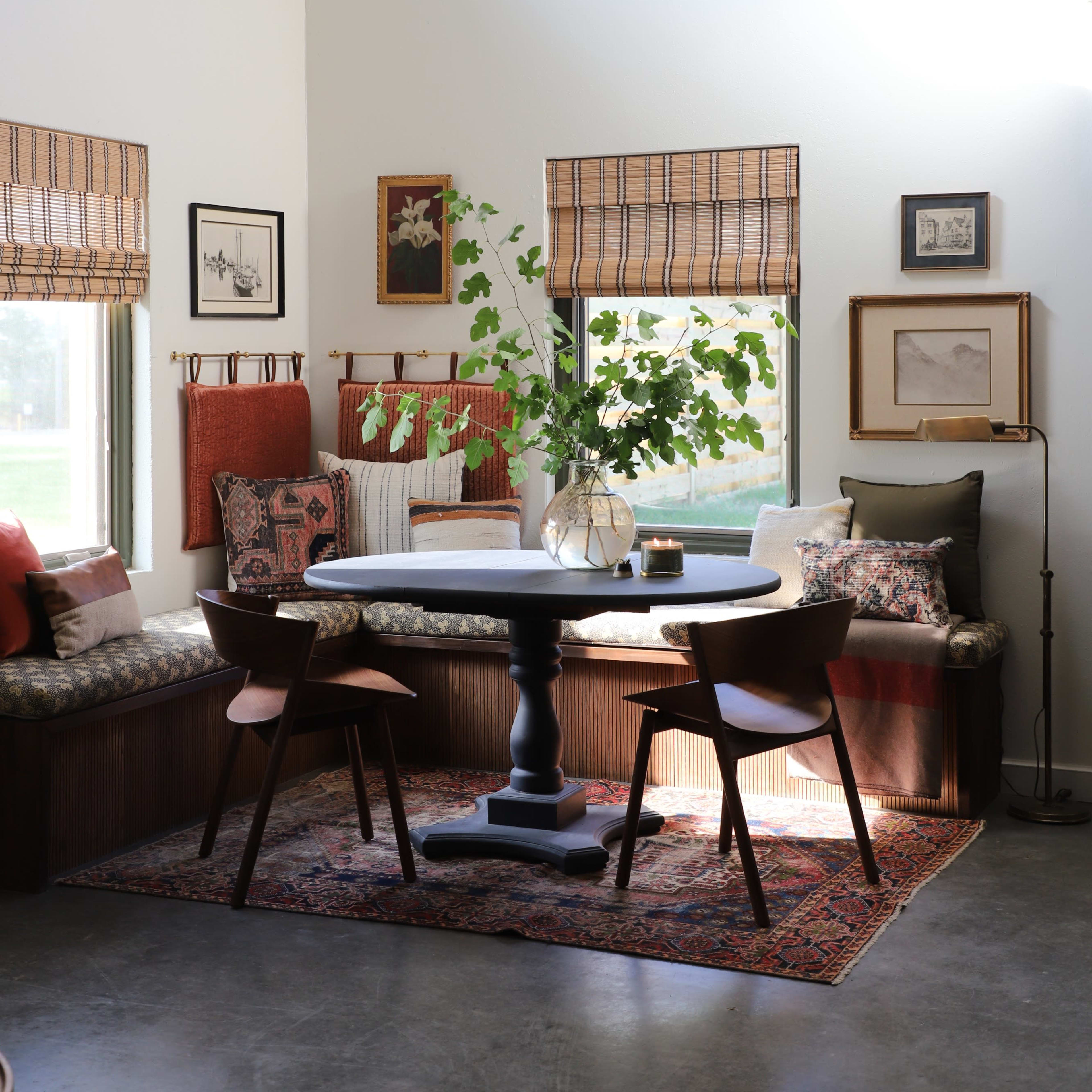 A cozy dining area features a round table with two chairs, surrounded by a window bench adorned with cushions and decorative art on the walls.