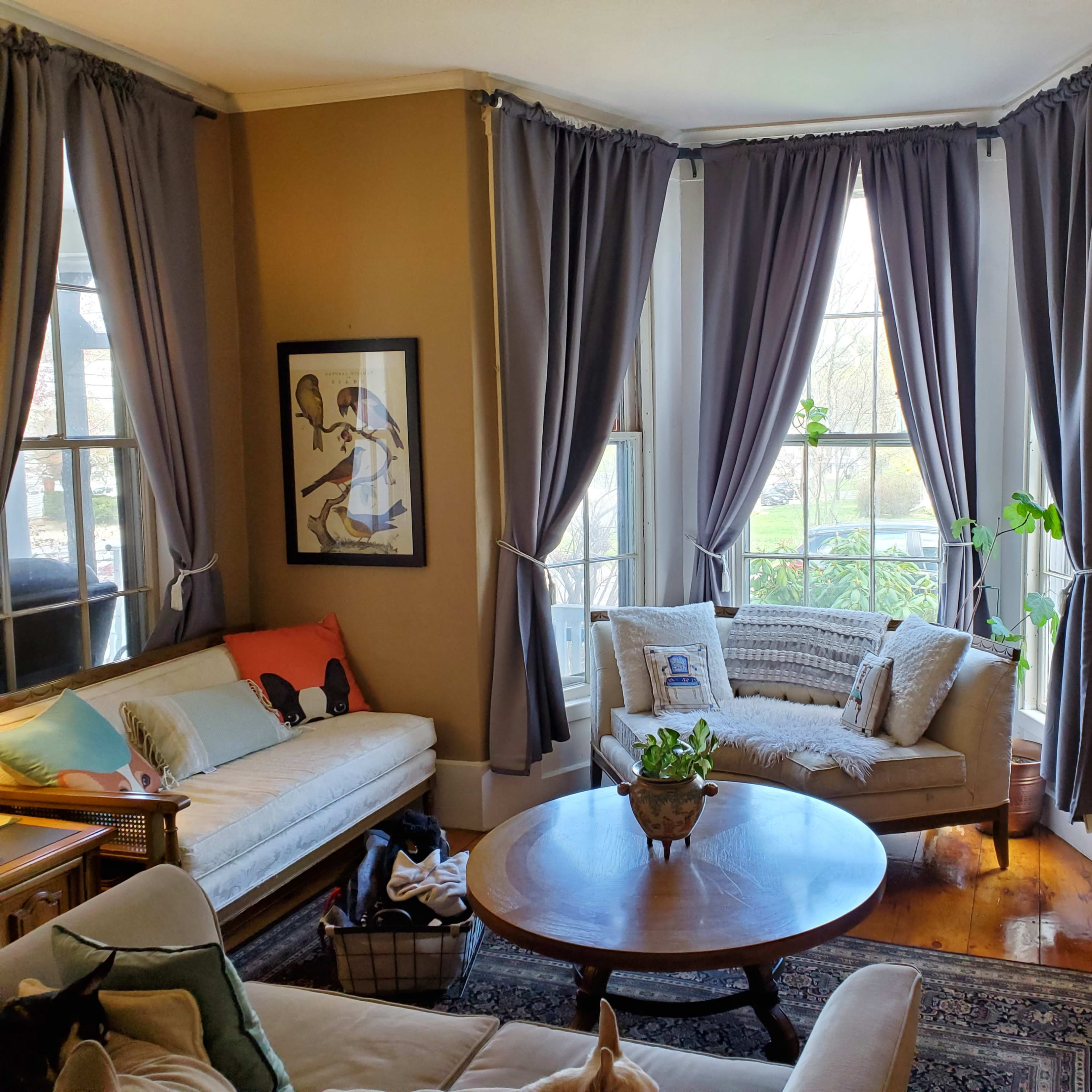 A living room features two sofas, a round coffee table, and large windows dressed with gray curtains, allowing natural light to fill the space.