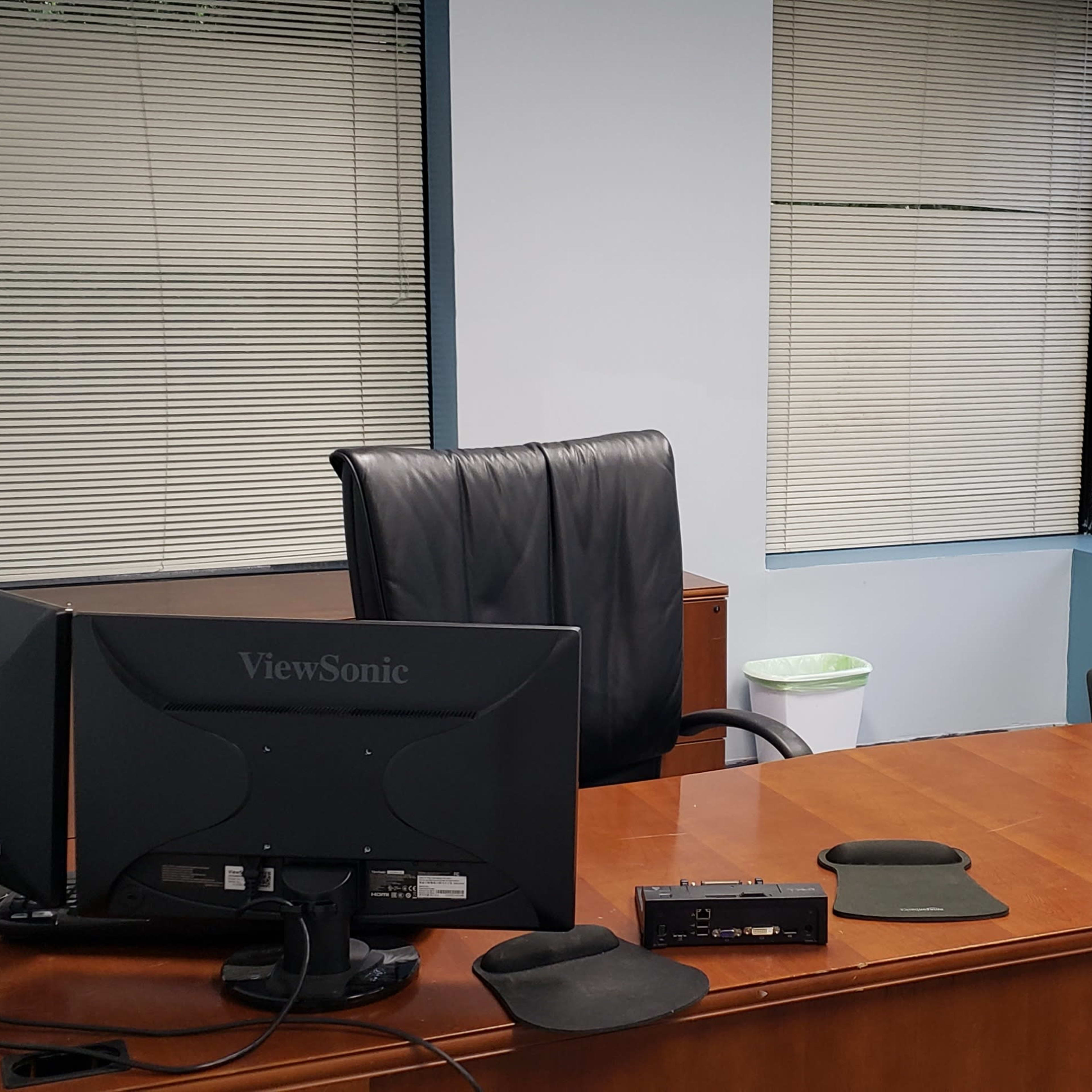 A small office with a wooden desk, an office chair, a computer monitor, and a green armchair beside two windows with blinds.