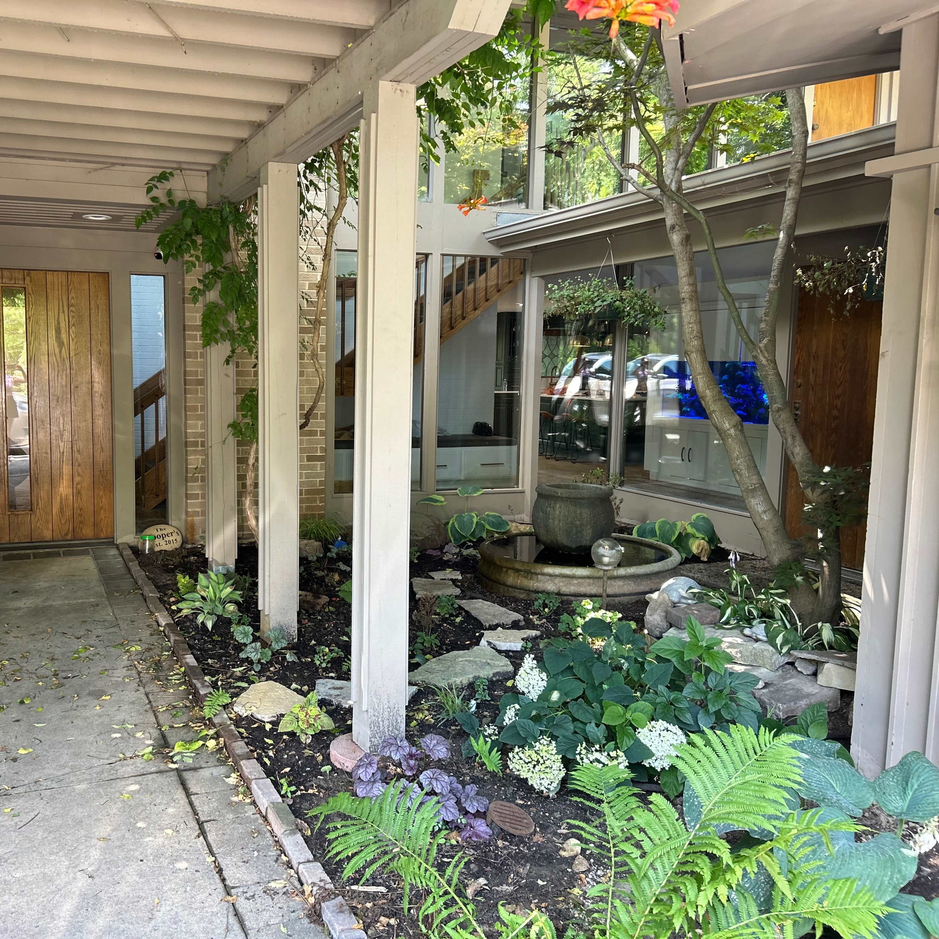 A landscaped entrance with stone pathways, plants, and a fountain, framed by wooden doors and a covered walkway.