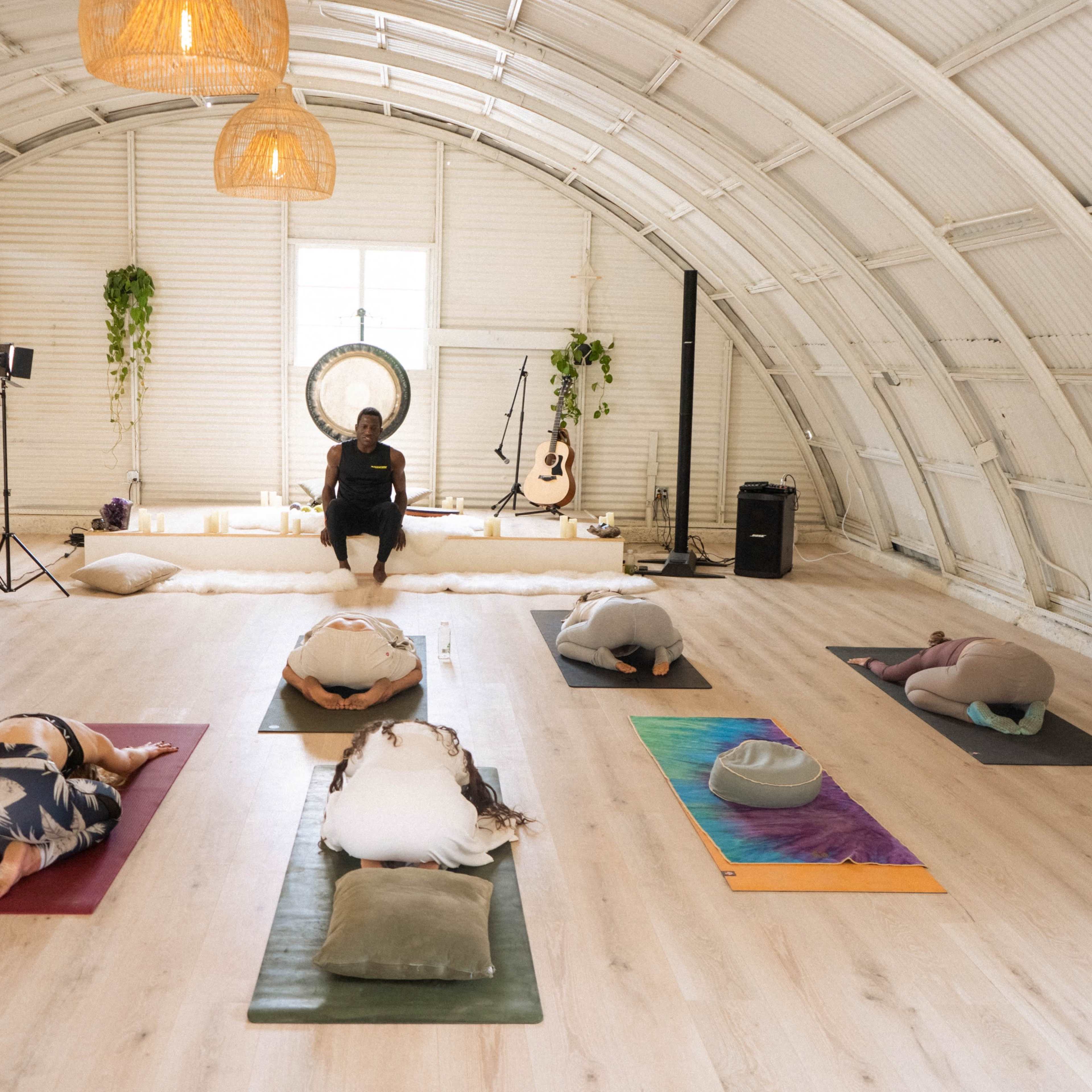 A group of individuals practice yoga in a spacious, airy studio while a leader sits in front of them.