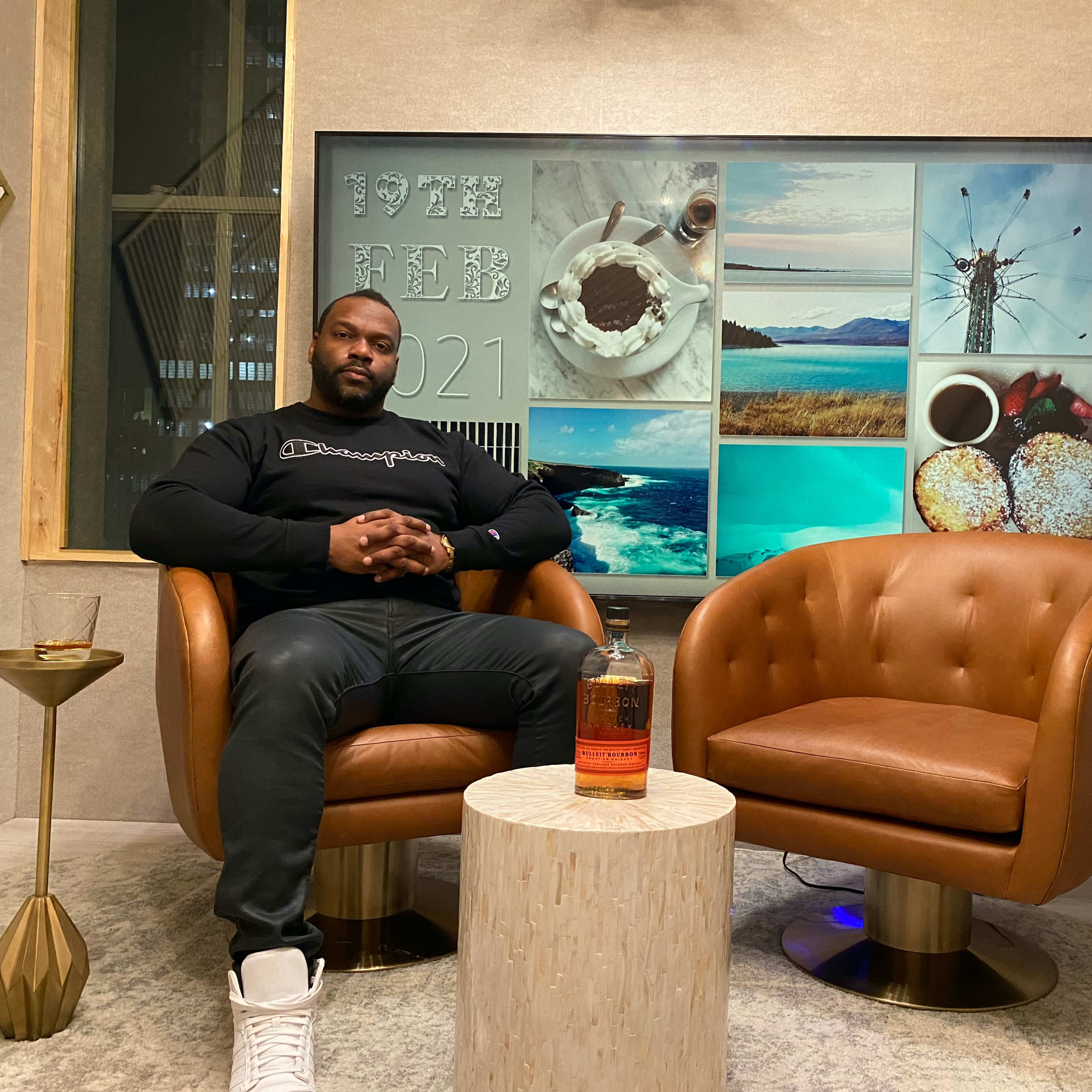A man sits between two brown lounge chairs, with a bottle of whiskey on a side table, against a backdrop of various scenic photographs and the date displayed on a wall.