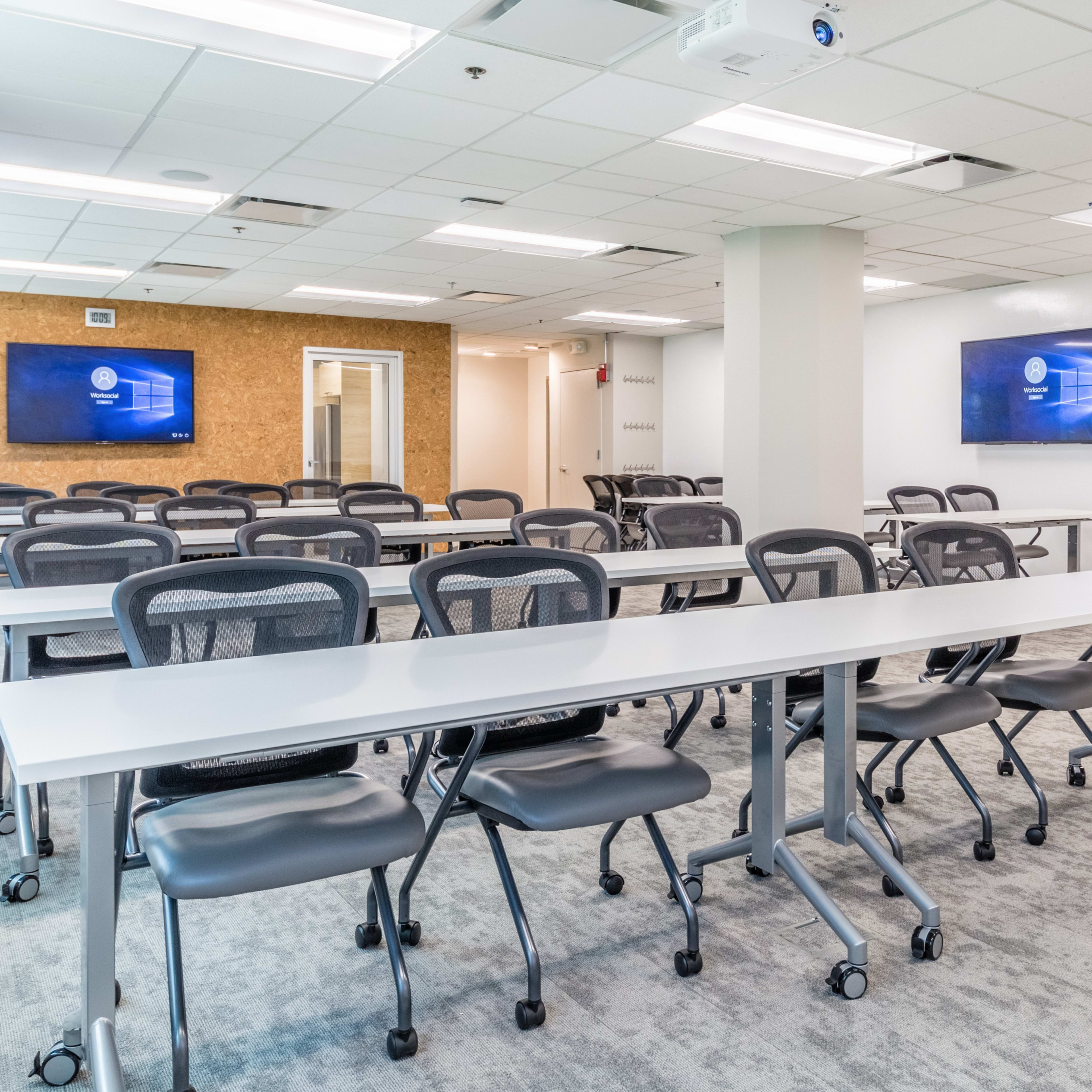 A modern classroom setup with multiple tables and chairs arranged in rows, featuring two wall-mounted screens.