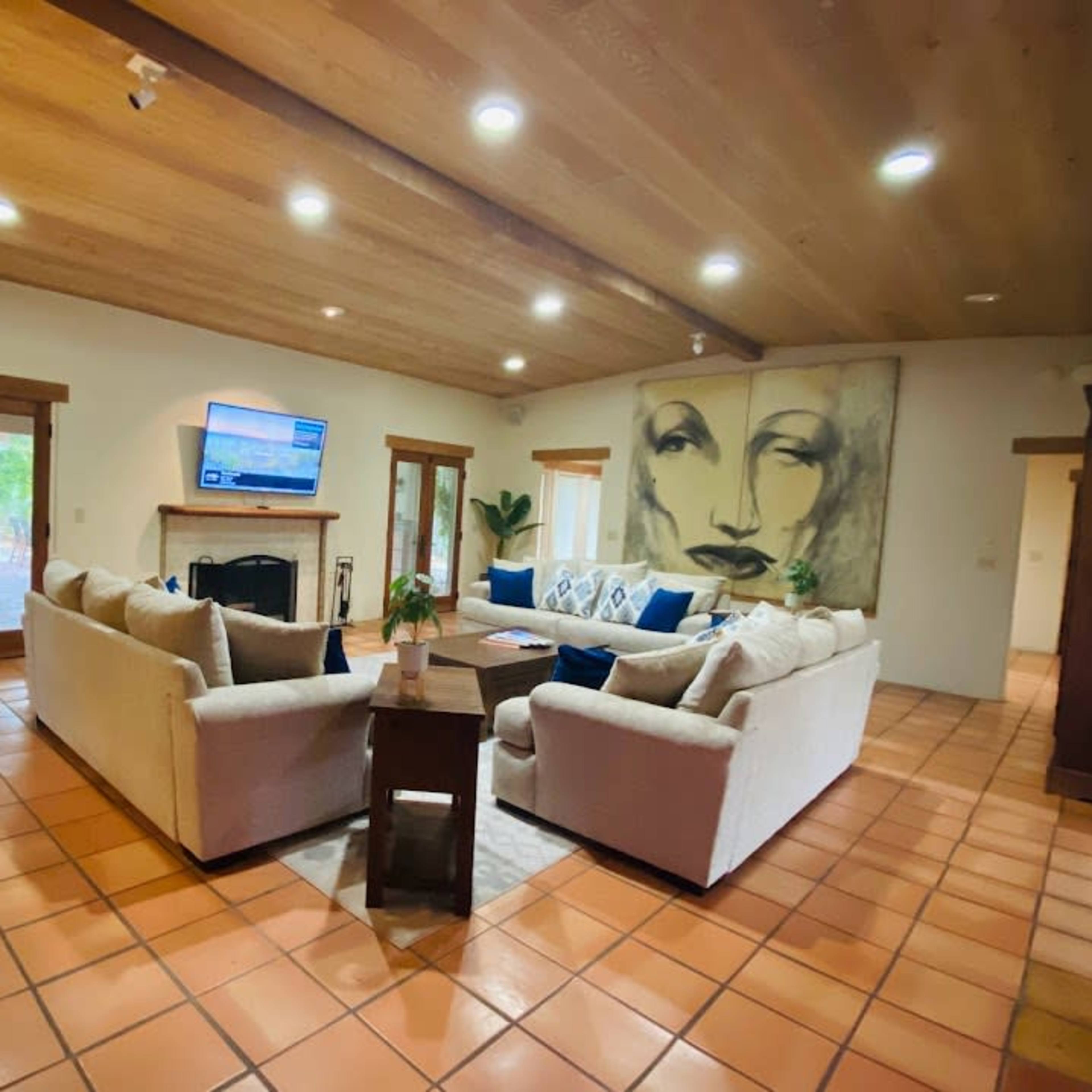 A spacious living room features a large sectional sofa, a wooden coffee table, and a flat-screen TV mounted on the wall, with a large artwork of a face and sliding glass doors leading to an outdoor area.