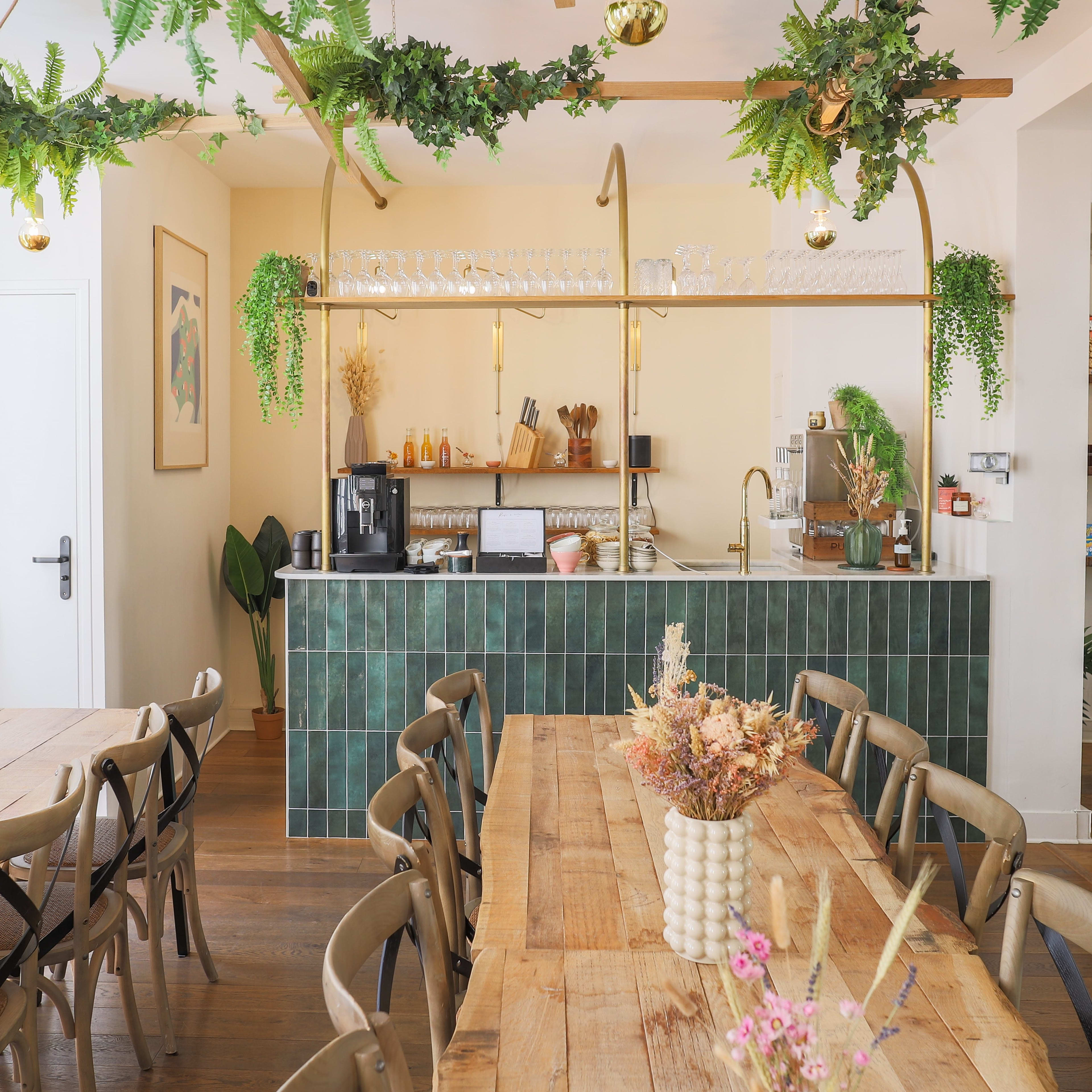 A modern café interior featuring a bar with green tile, wooden tables, and decorative plants hanging from the ceiling.